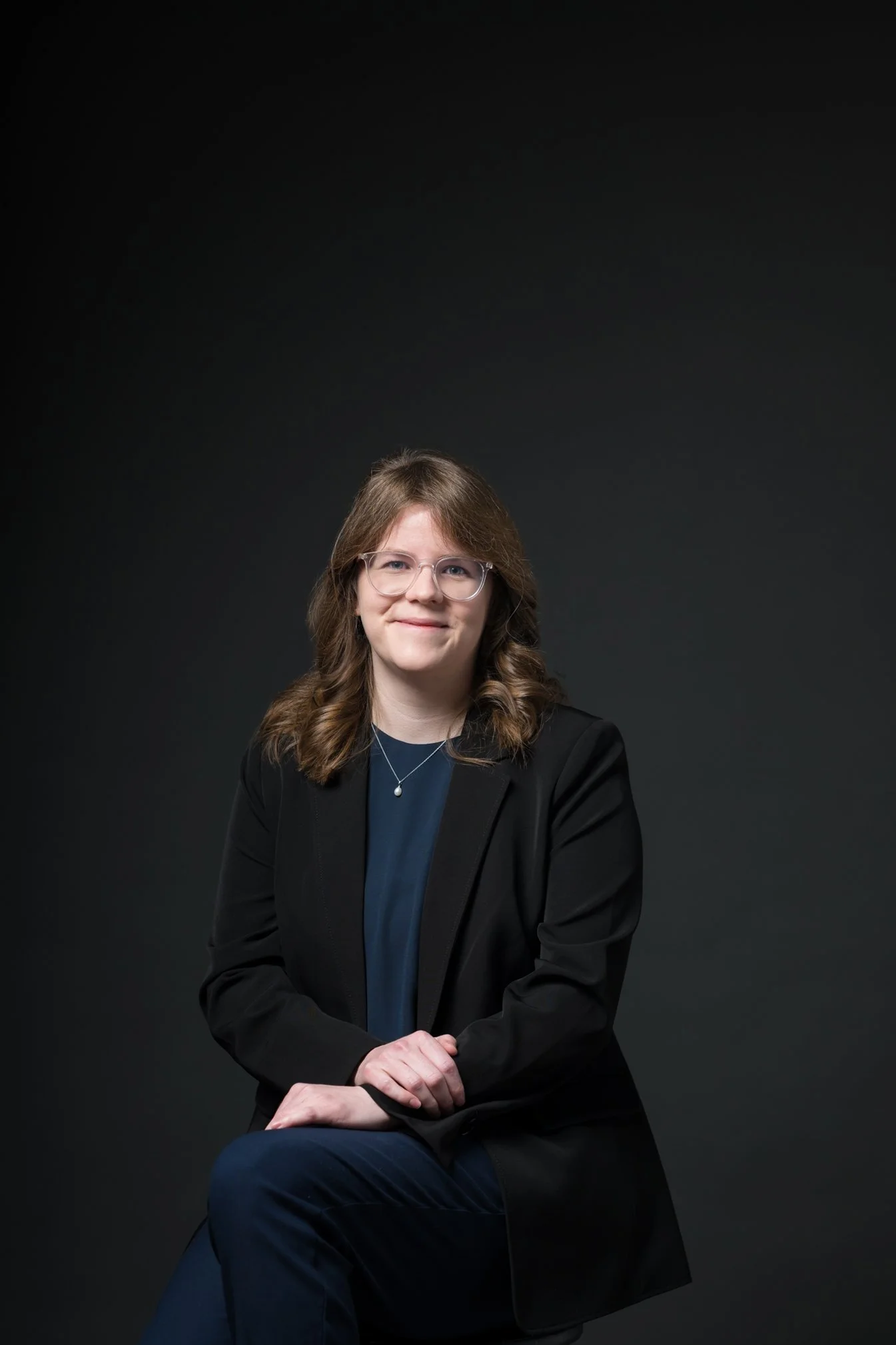 Woman with curly brown hair and glasses wearing a black blazer and patterned blouse sitting against a dark background.
