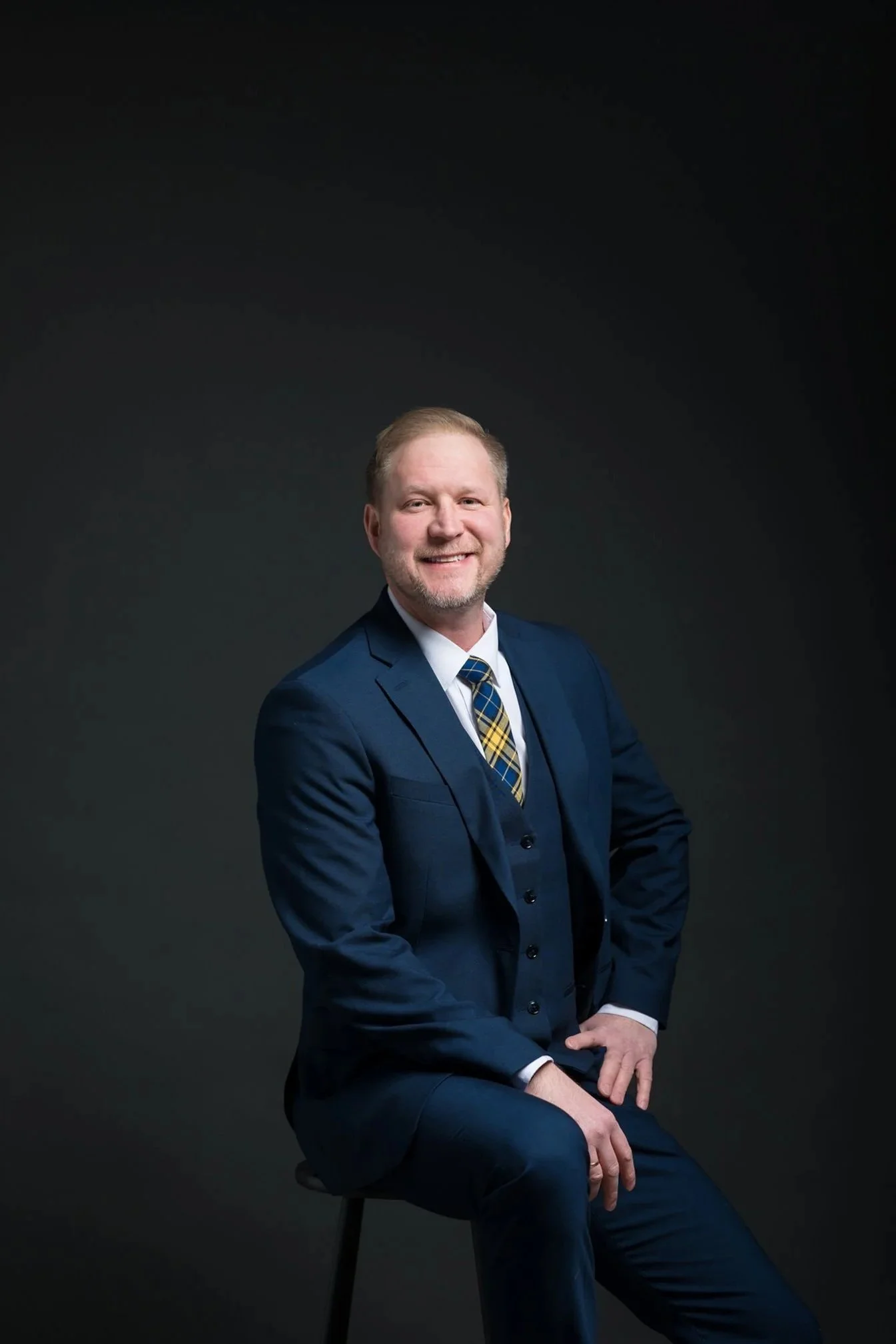 A man in a navy blue suit with a plaid tie sitting on a stool against a dark background, smiling at the camera.