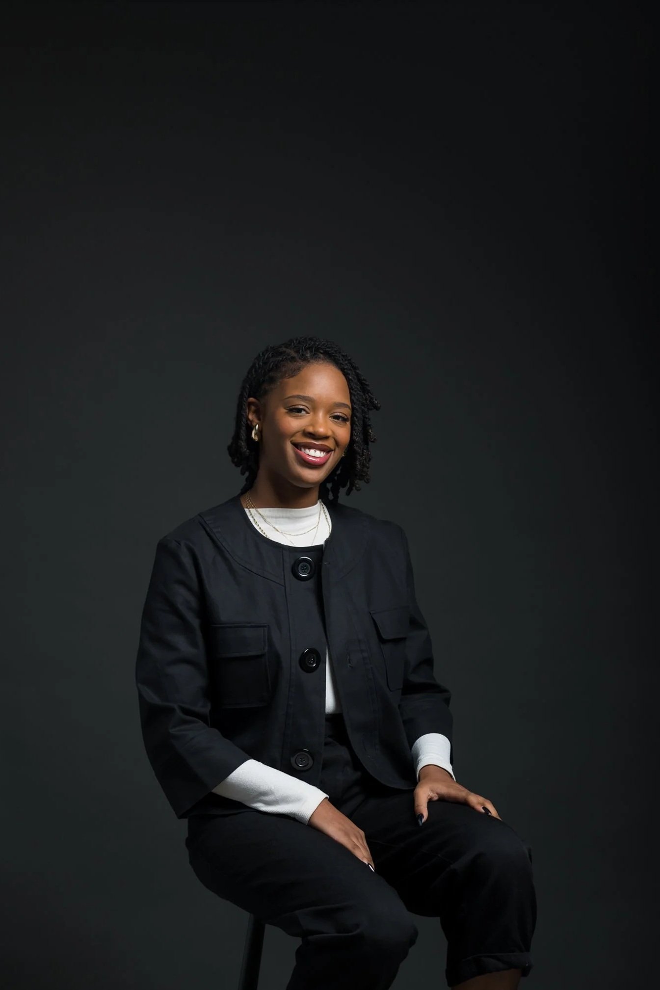 A smiling woman with natural curly hair, sitting on a stool against a dark grey background, wearing a black jacket and white shirt.