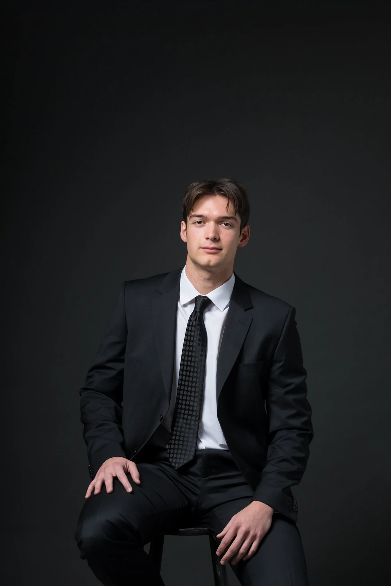 A young man in a black suit sitting on a stool against a dark background.