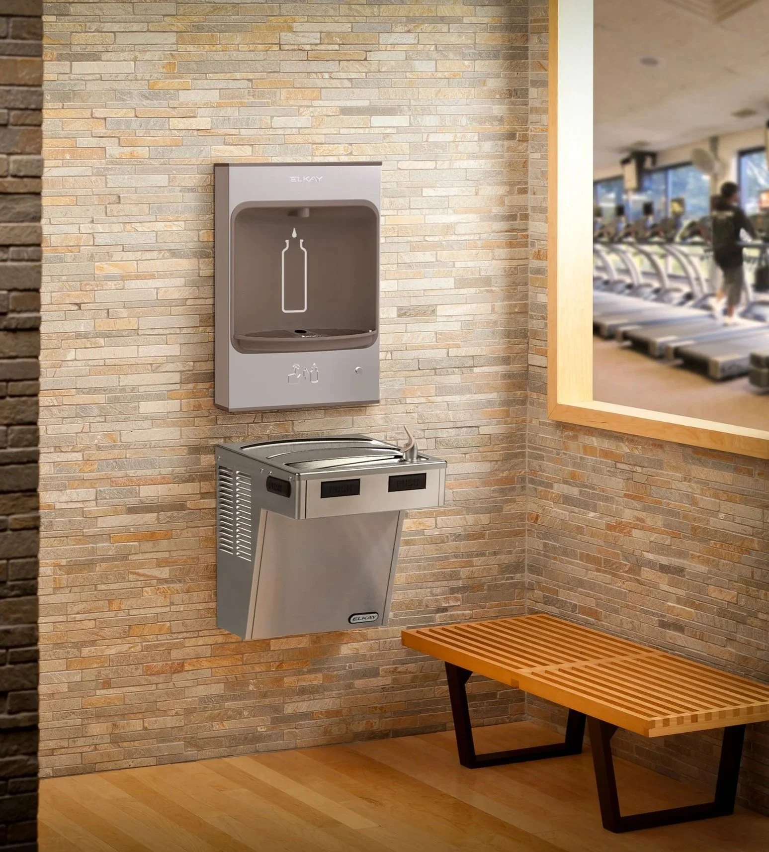 A water bottle filling station and a stainless steel drinking fountain in a gym lobby, with a wooden bench against the wall and a large window showing people exercising on treadmills inside.