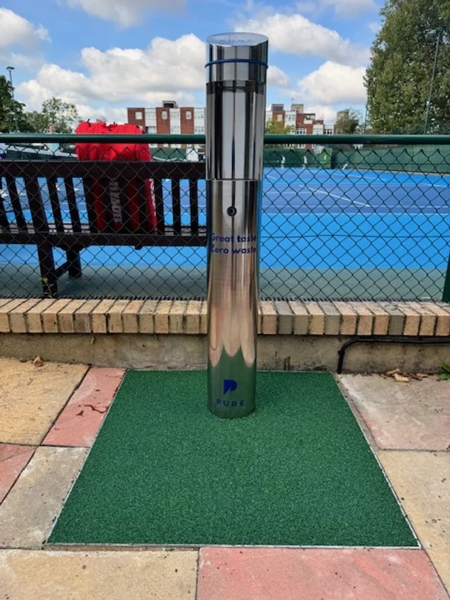 A stainless steel water refill station with a clear cap, installed on a small green mat outside near a tennis court, with a bench and trees in the background.