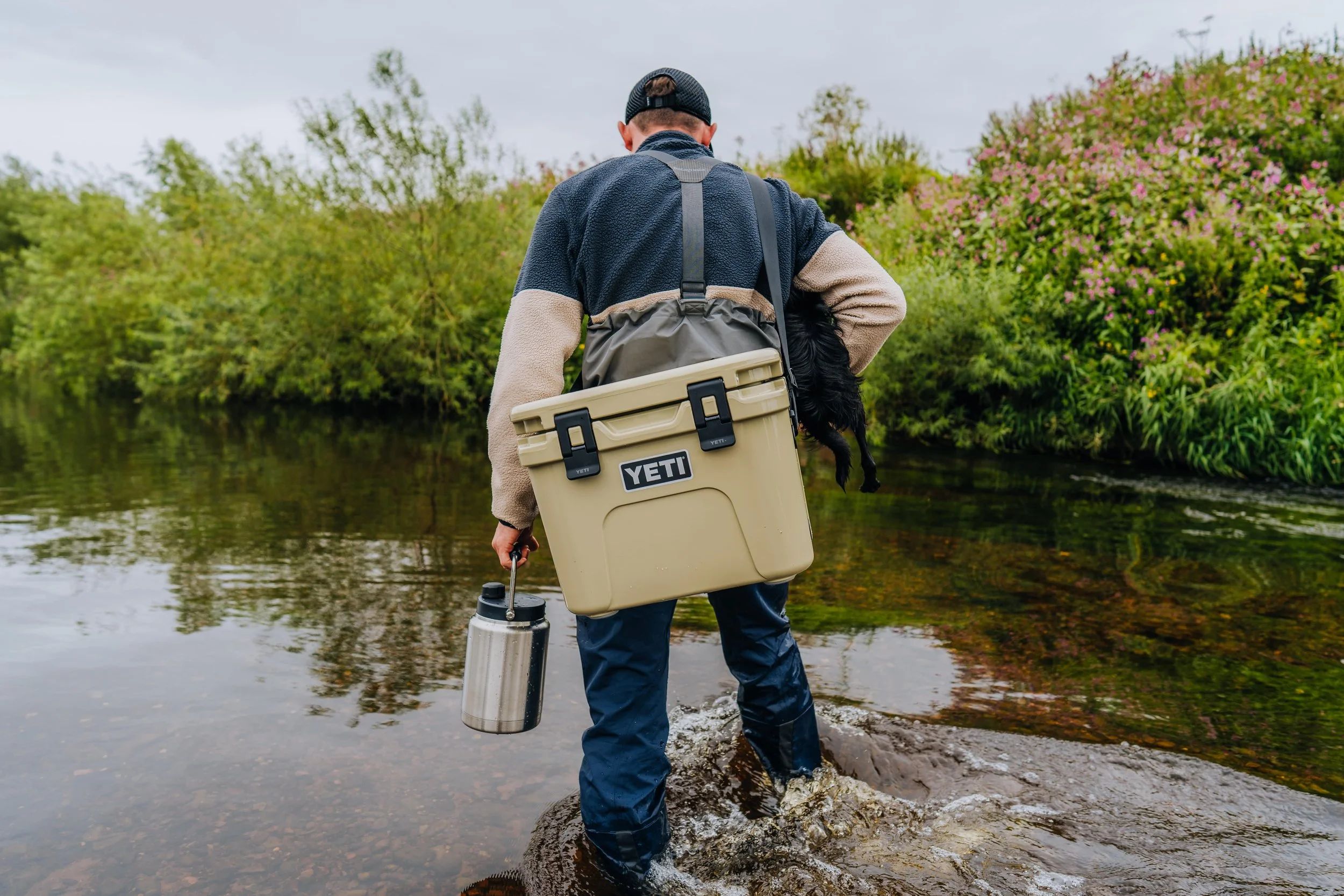Man wading through a creek carrying a YETI cooler, a stainless steel water bottle, and a small black dog in a harness, with lush green bushes and pink-flowering plants in the background.