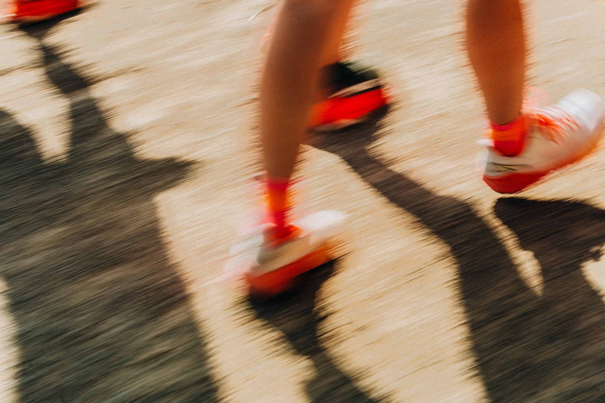 Blurred motion shot of a person running on a light-colored outdoor track, wearing white athletic shoes with red accents, red socks, and black shorts.