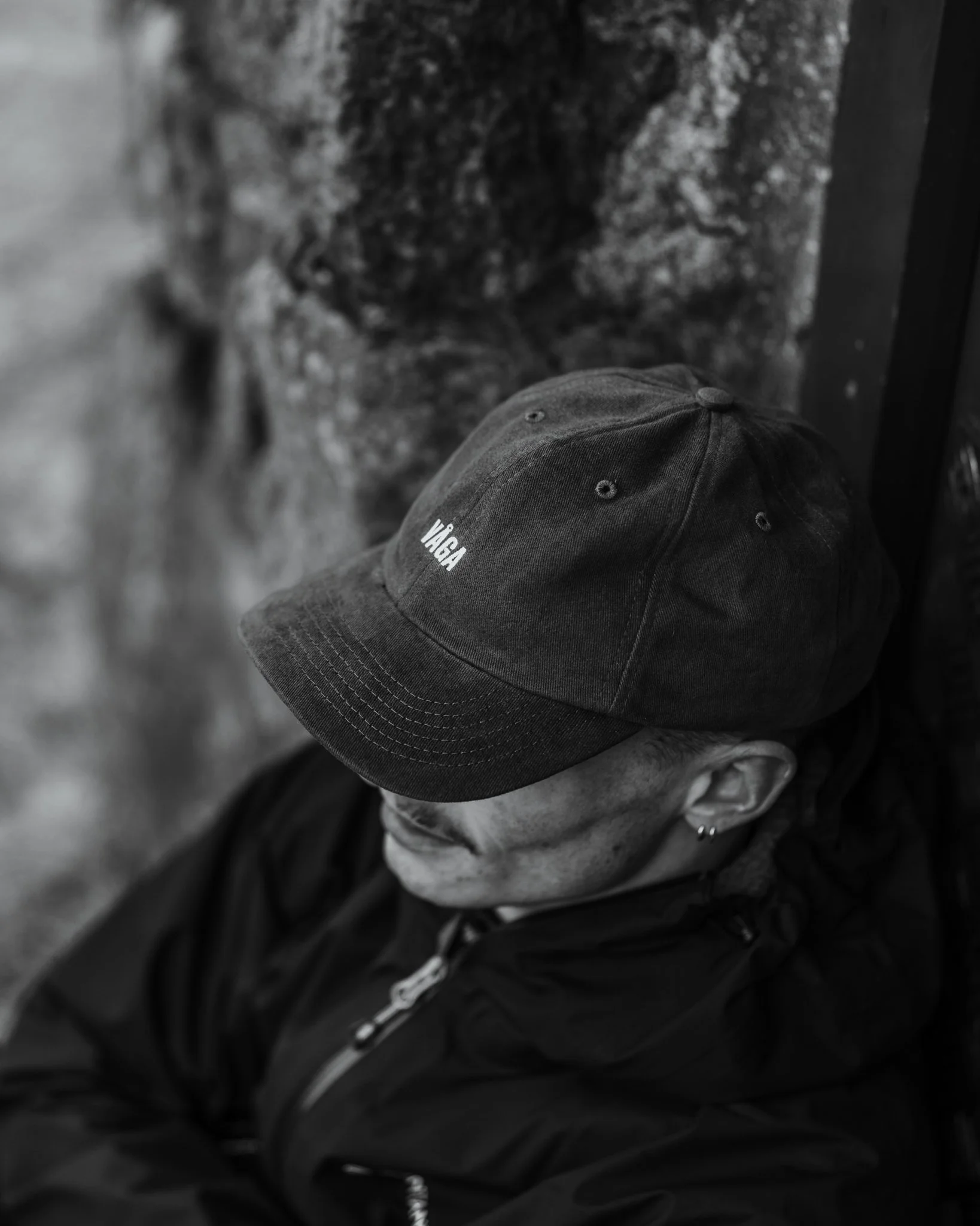 Black and white photo of a person wearing a cap with 'VIGA' written on it, leaning against a stone wall, with their face partially visible and turned downward.