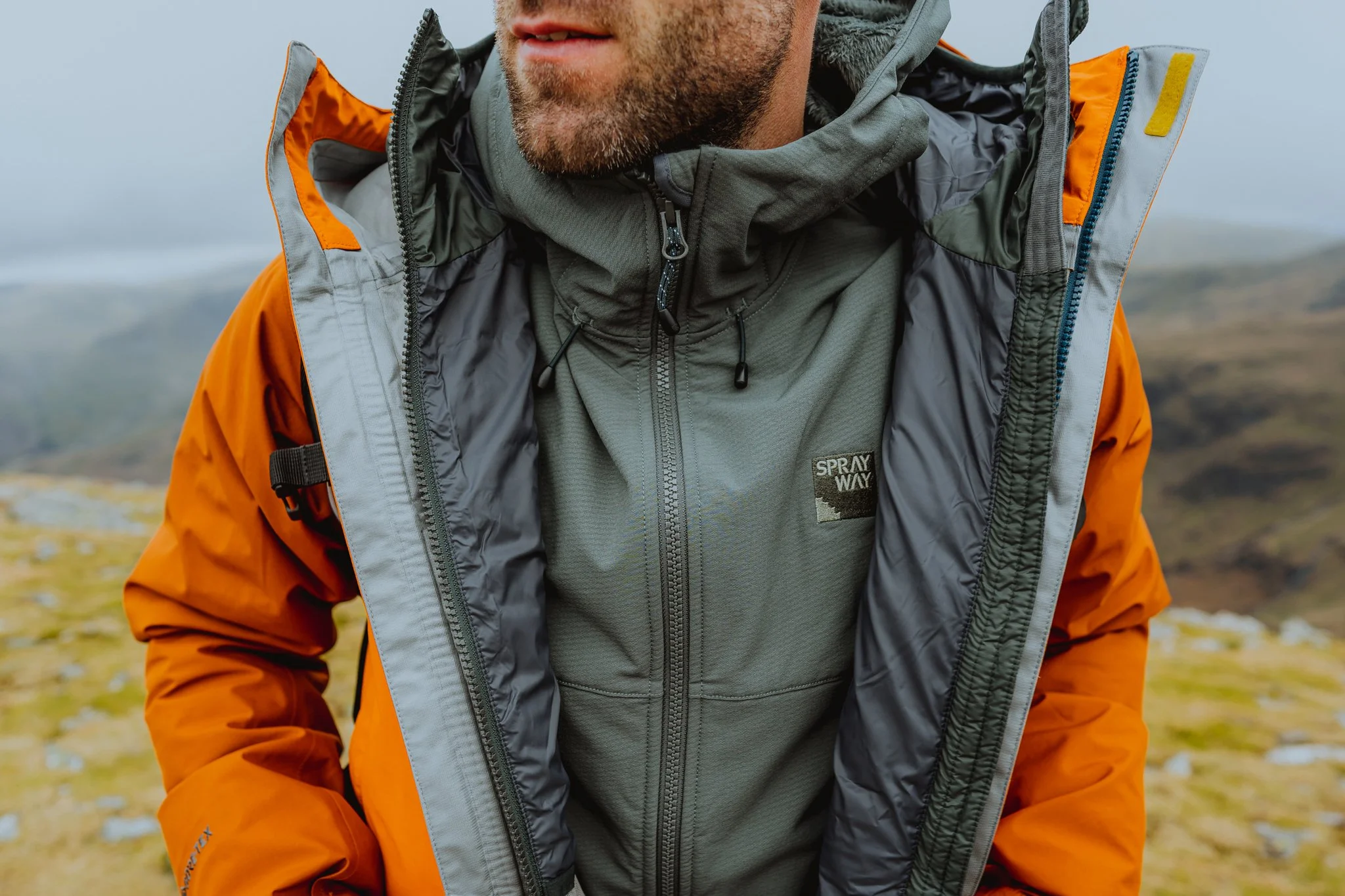 Freelance Lifestyle Photographer  Close-up of a man wearing an orange and grey outdoor jacket and a grey fleece jacket with a mountain landscape in the background.