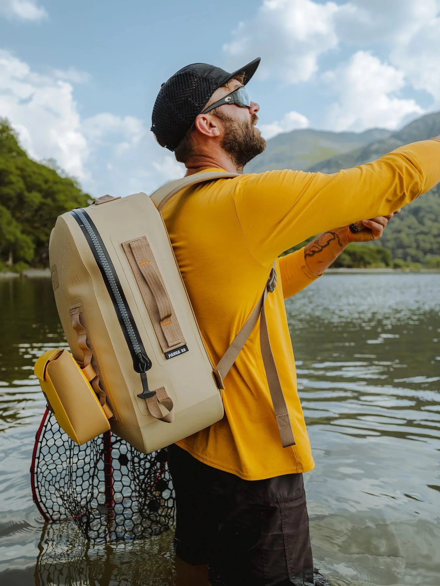 A man in a yellow jacket and black hat with sunglasses stands in a body of water, reaching out with his right arm while carrying a beige backpack with a yellow attached pouch. There are green trees on the left side and a mountain in the background.