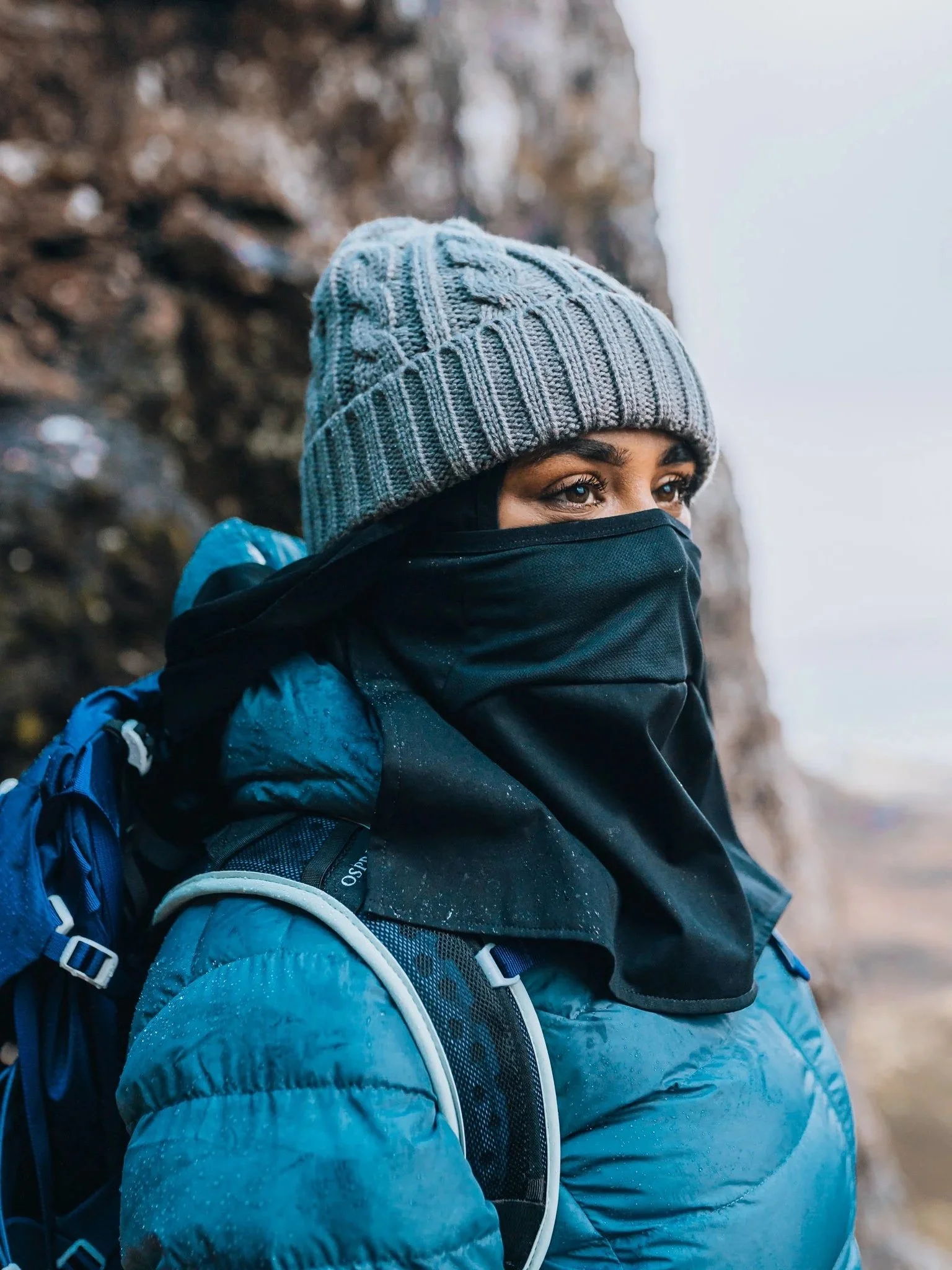 Woman wearing a gray knit beanie, black face covering, and blue jacket with a backpack outdoors.