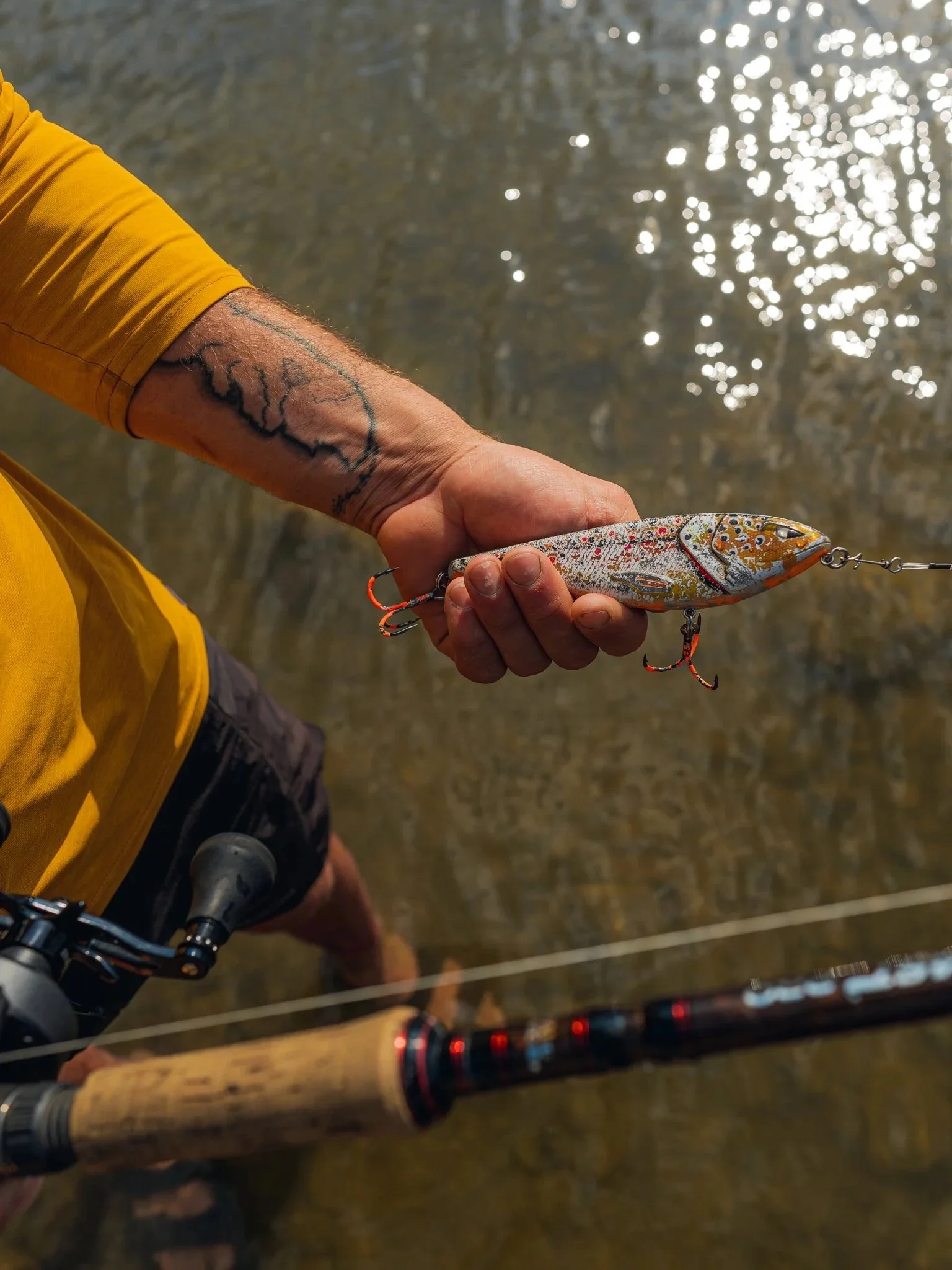 Person standing in shallow water holding a colorful fishing lure with a fishing rod in the foreground.