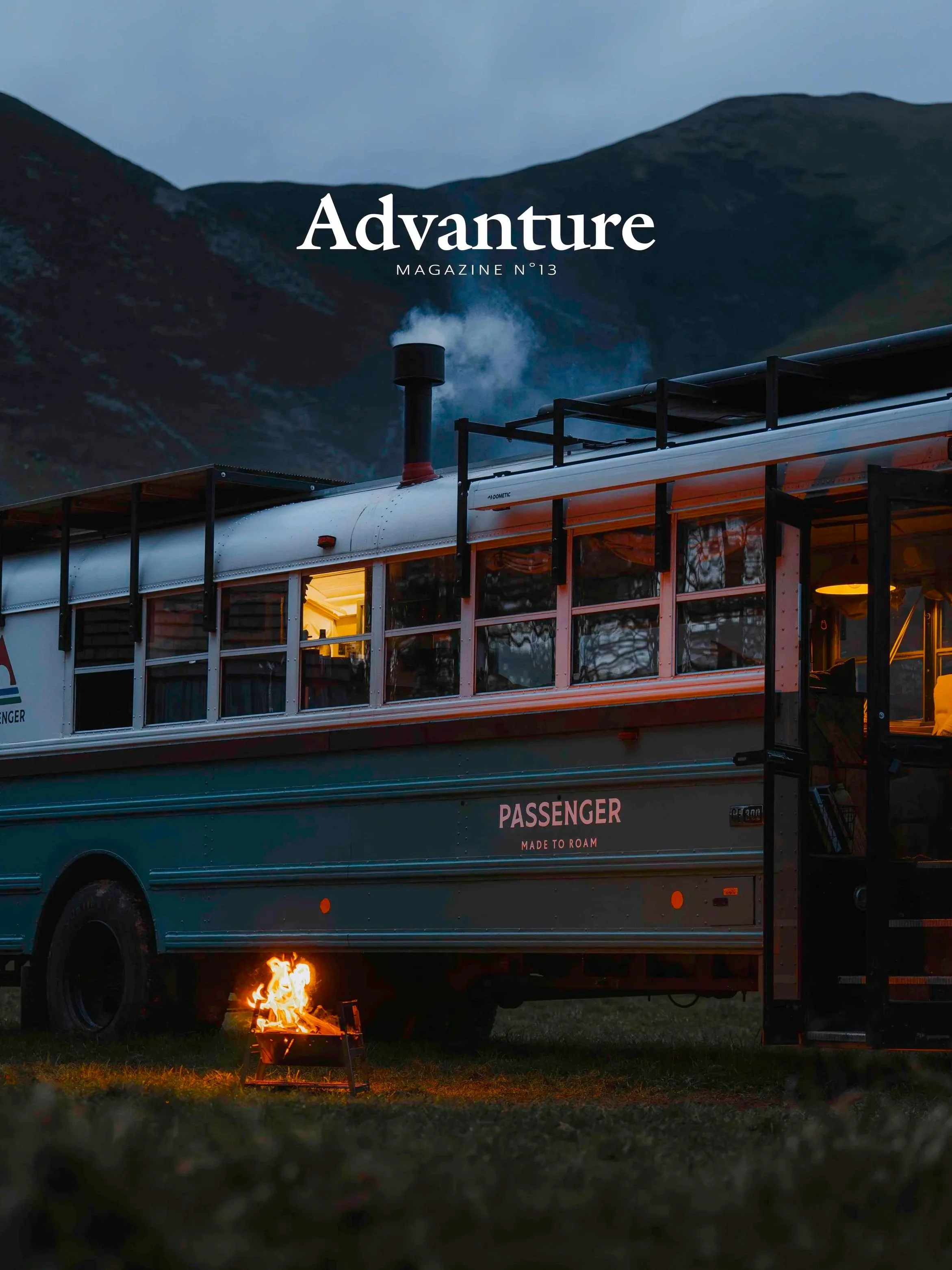 A vintage bus with open windows parked in a grassy field during dusk, with a small campfire burning in front, mountains in the background, and a darkening sky.