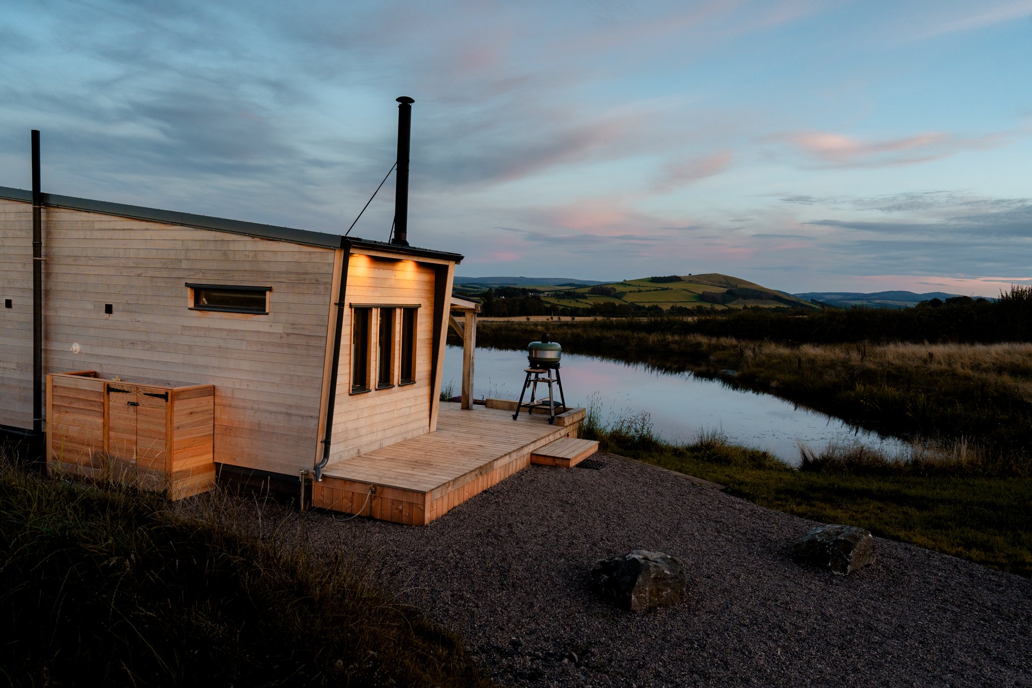 Freelance outdoor Lifestyle Photographer A modern wooden cabin with a small deck overlooking a river at sunset, surrounded by grassy fields and distant hills.