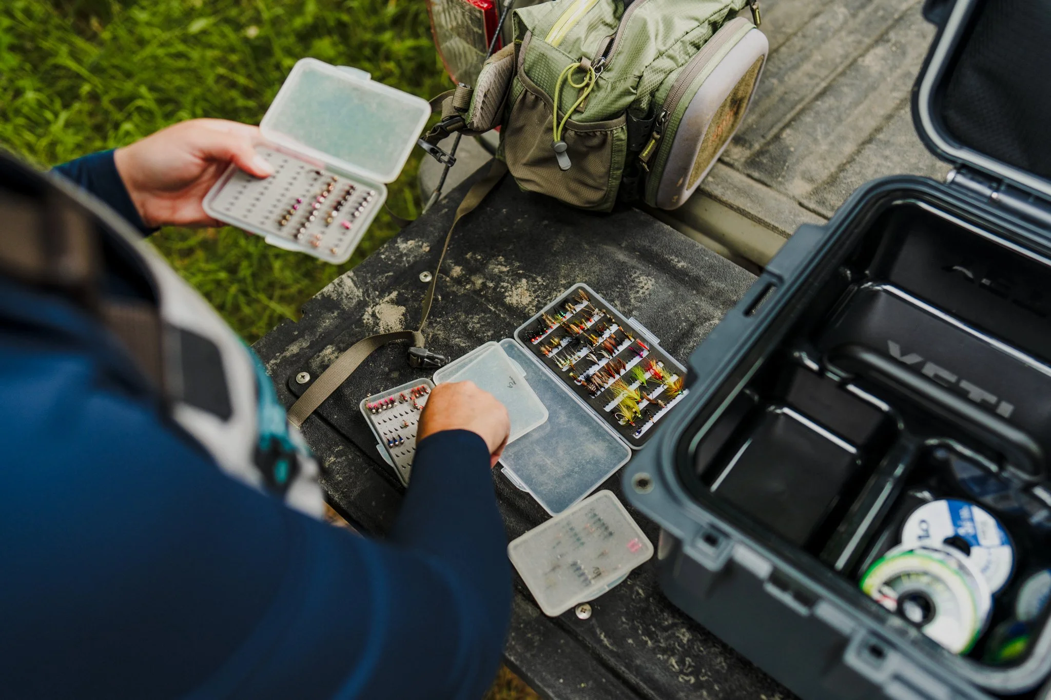 Freelance Lifestyle Photographer Person organizing fishing lures and tackle boxes on a weathered outdoor table, with fishing gear and a backpack nearby.