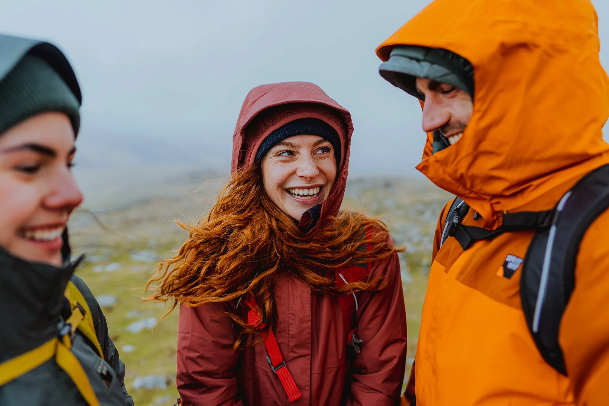 Three friends outdoors in jackets and hats, smiling and talking together in a mountainous area on a cloudy day.