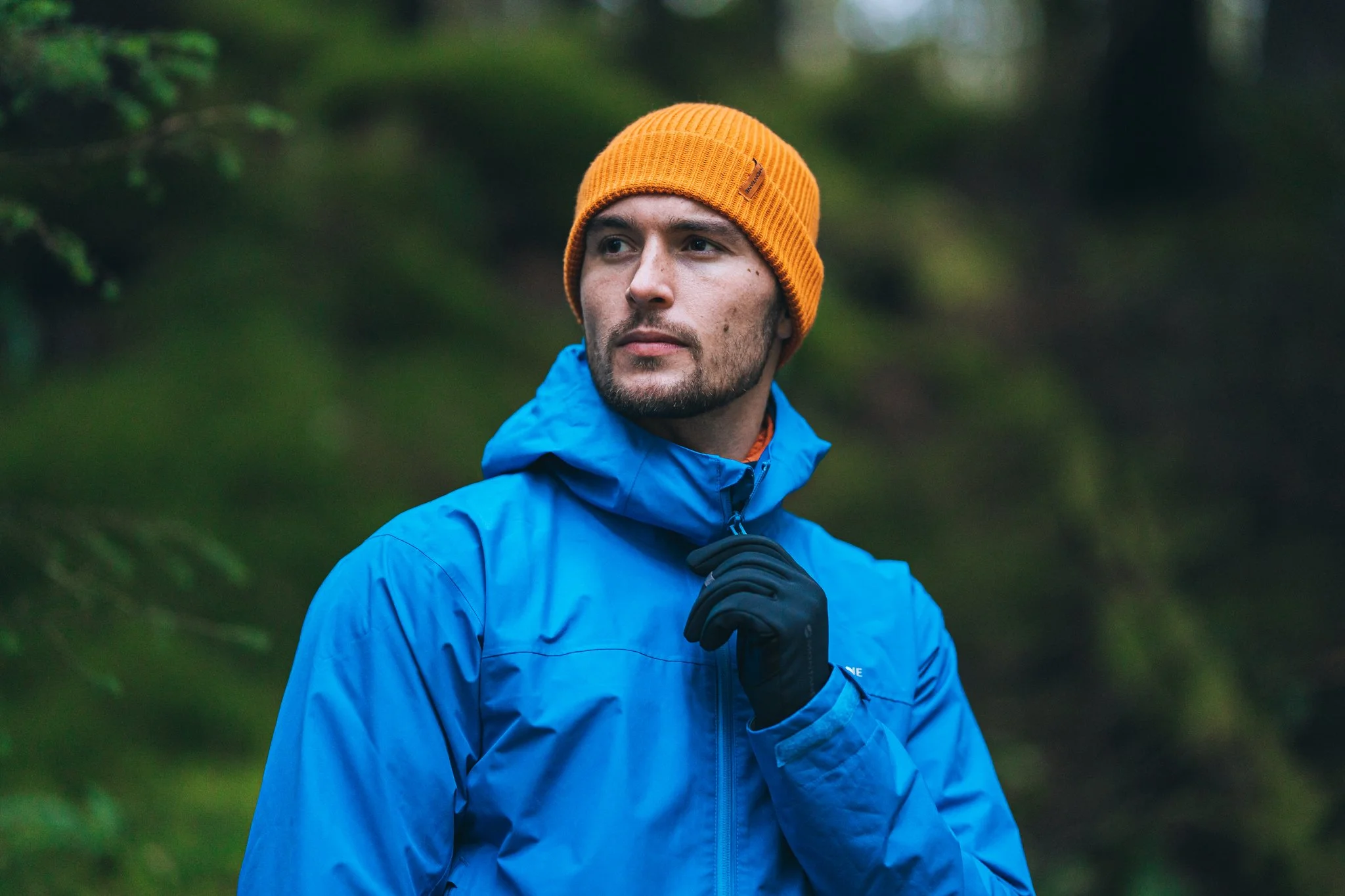 Man outdoors wearing a blue jacket, orange beanie, and black gloves, looking to the side in a green forest setting.