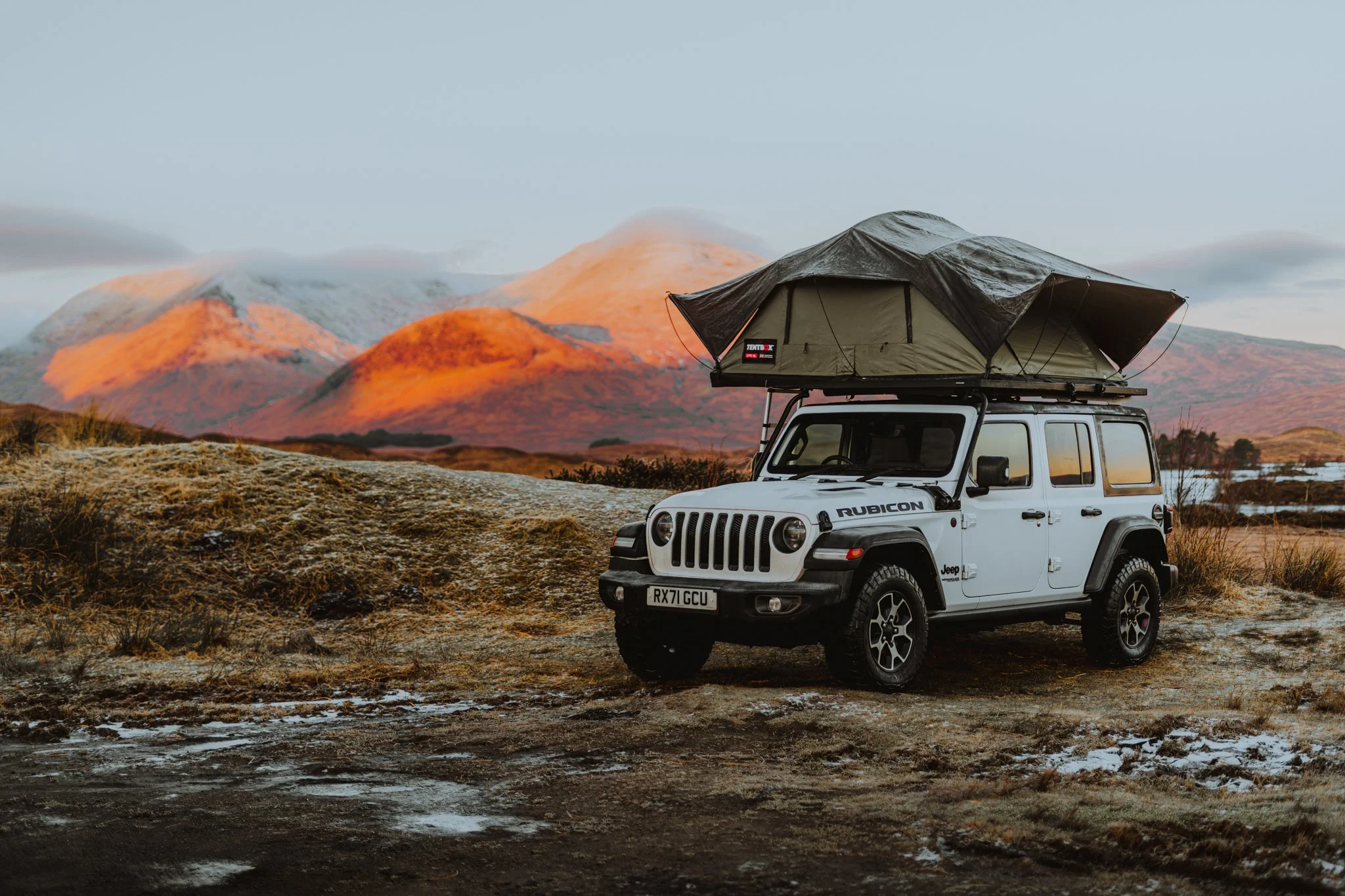 A white Jeep Rubicon with a rooftop tent in a rugged landscape with mountains in the background.