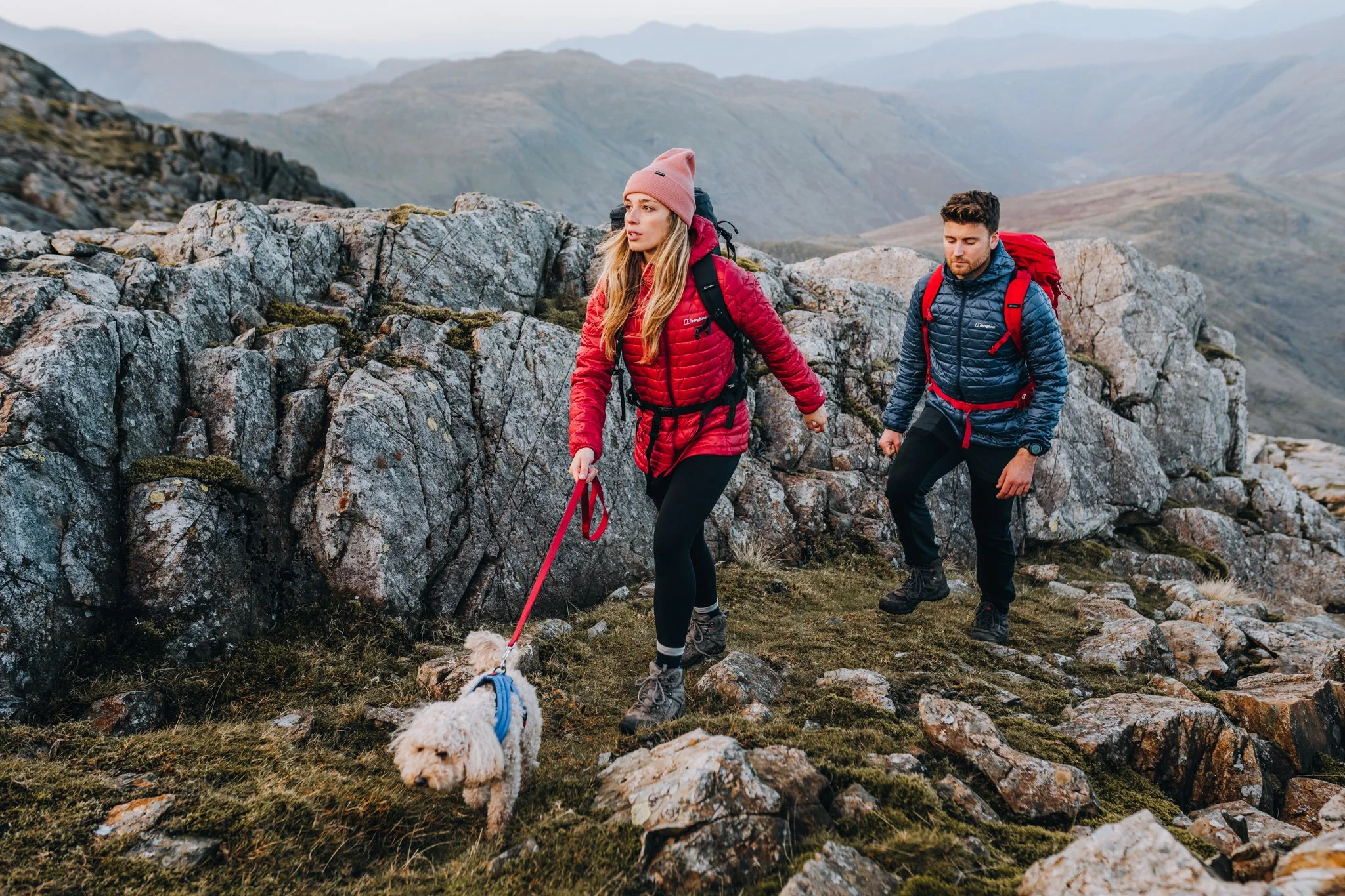 Two hikers, a woman and a man, climbing over rocky terrain in a mountainous landscape with a small white dog on a leash.