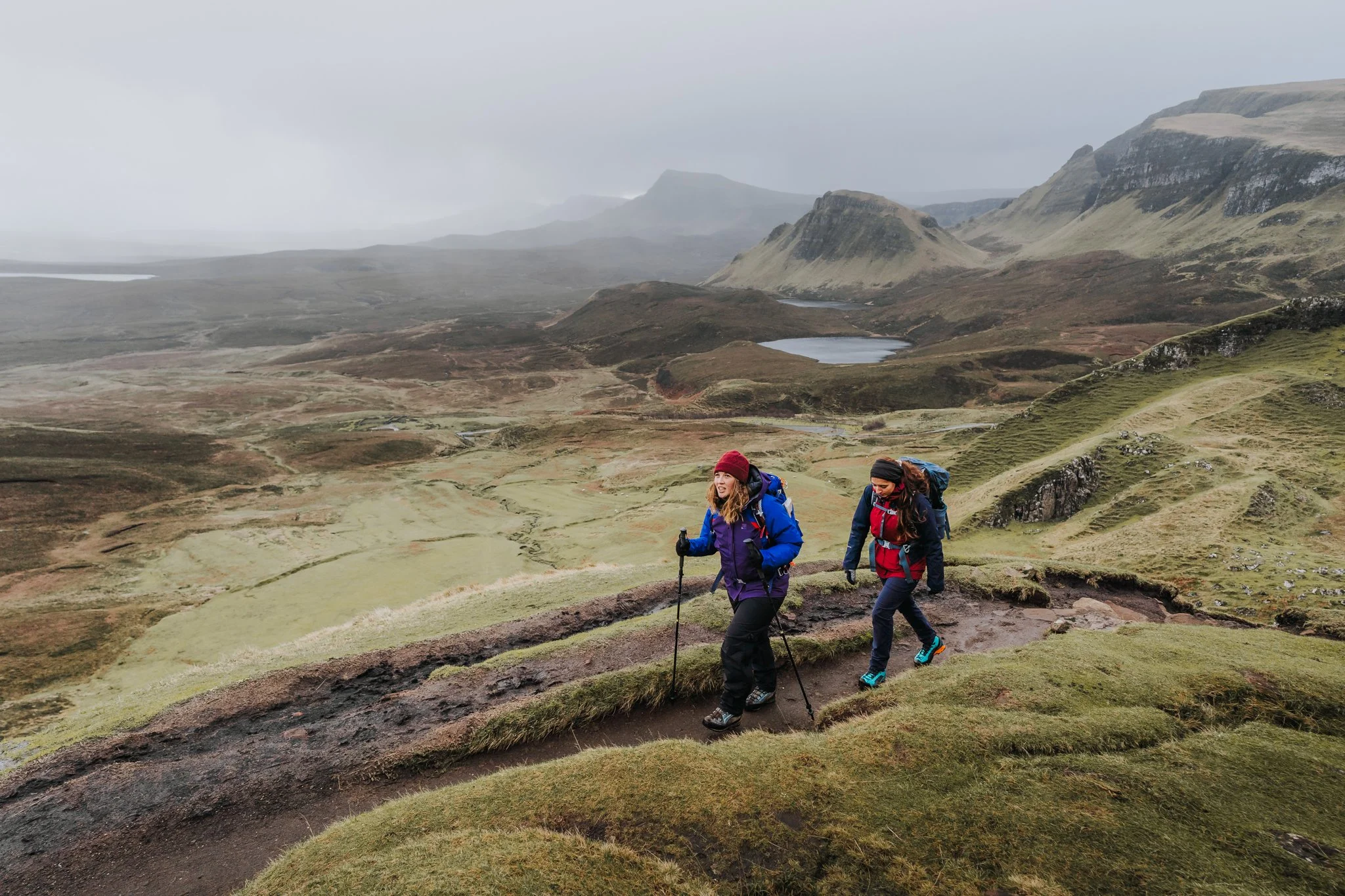 Two hikers, dressed in outdoor gear, trek along a grassy trail in a remote, rugged landscape with rolling hills, mountains, a pond, and a cloudy sky in the background.