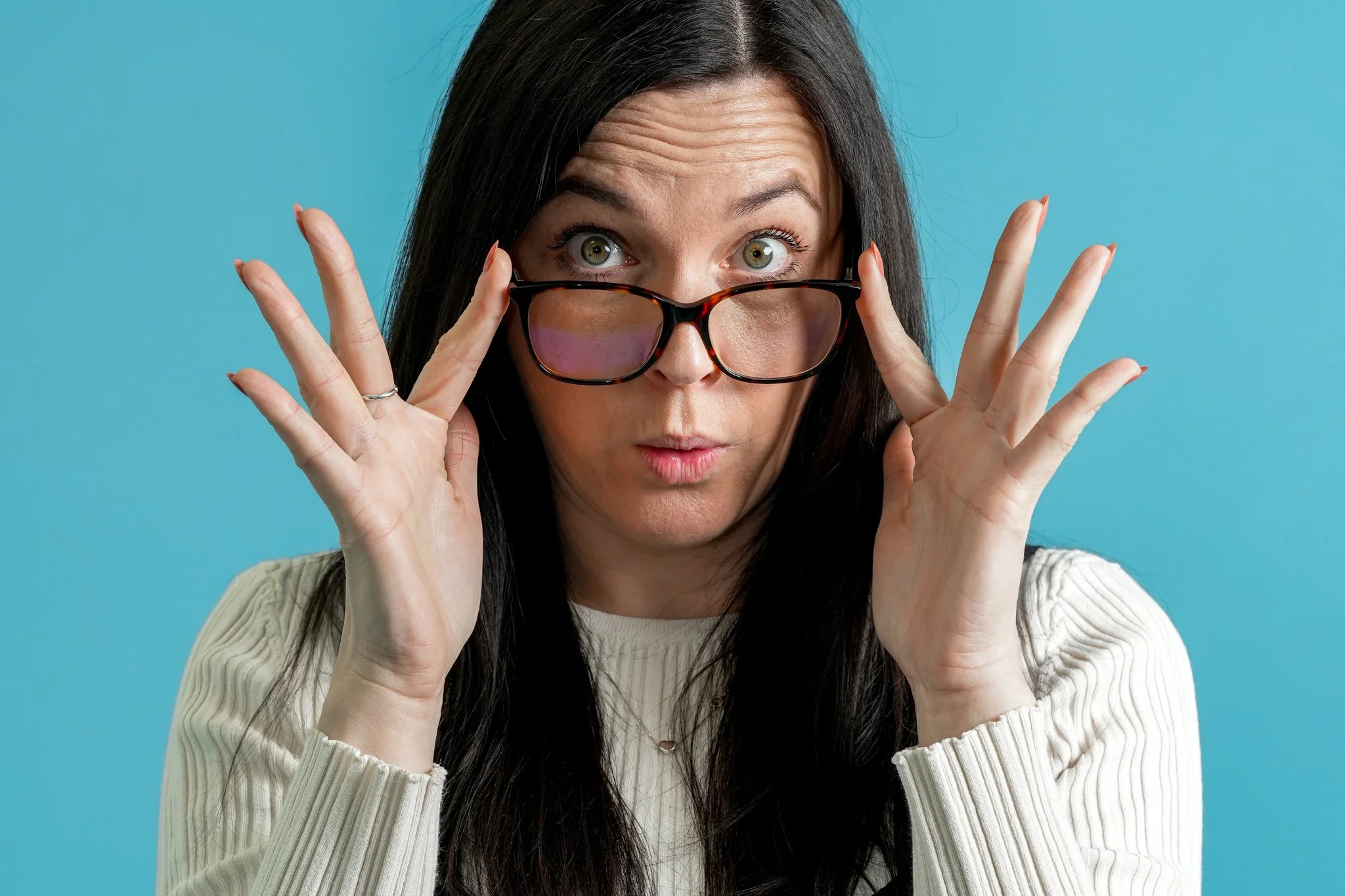 Freelance Lifestyle Photographer  Woman with dark hair, wearing glasses, pulling them down to look surprised or curious, against a blue background.