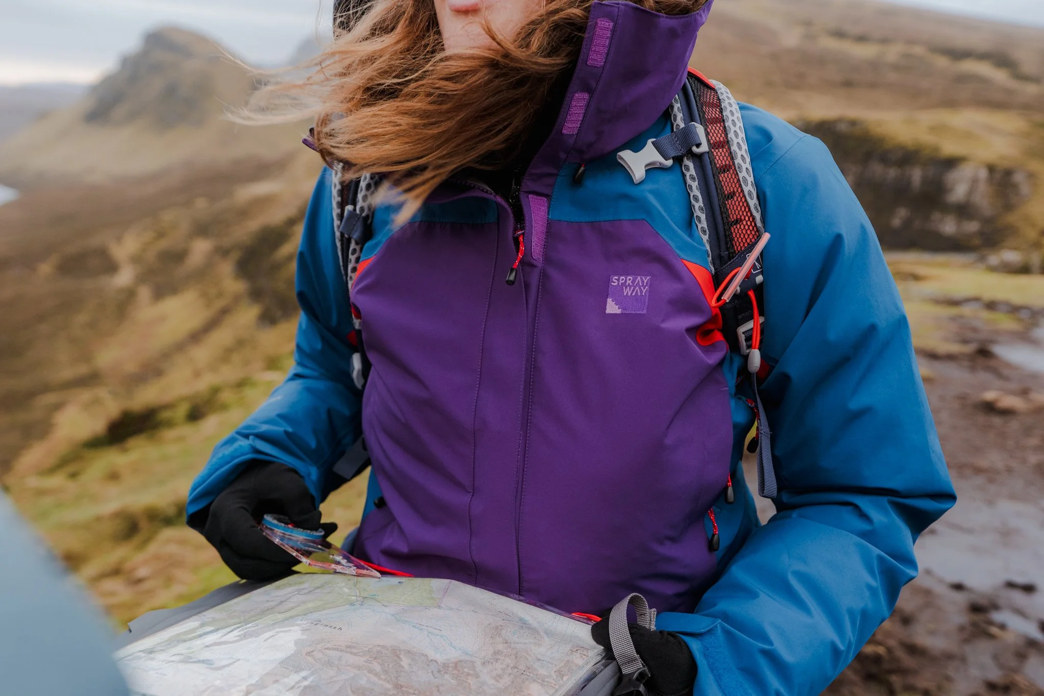 A person wearing a purple and blue outdoor jacket and black gloves holding a map outdoors with mountains in the background.