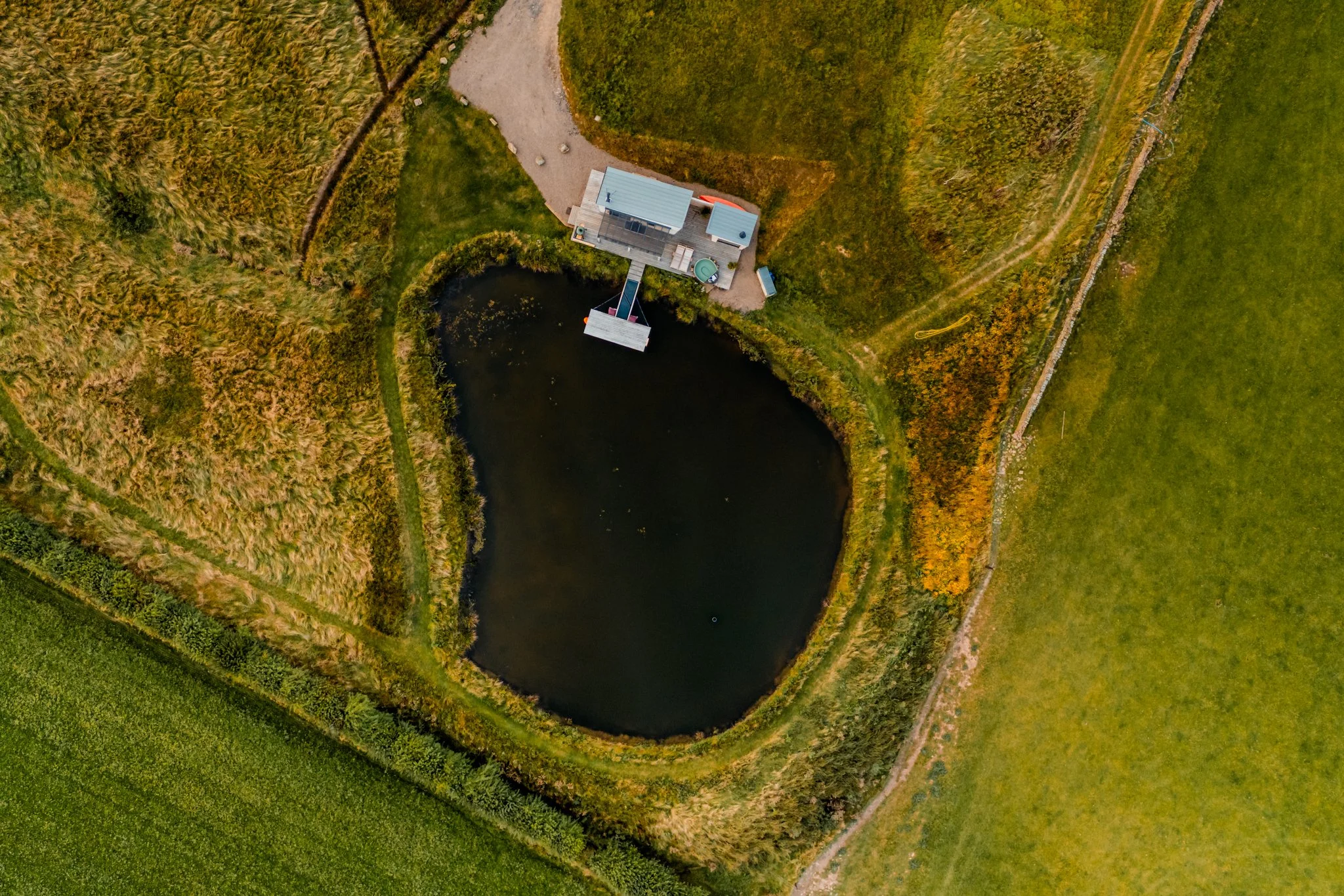 Aerial view of a small pond with a building and a dock on the shore, surrounded by green fields and rural landscape.