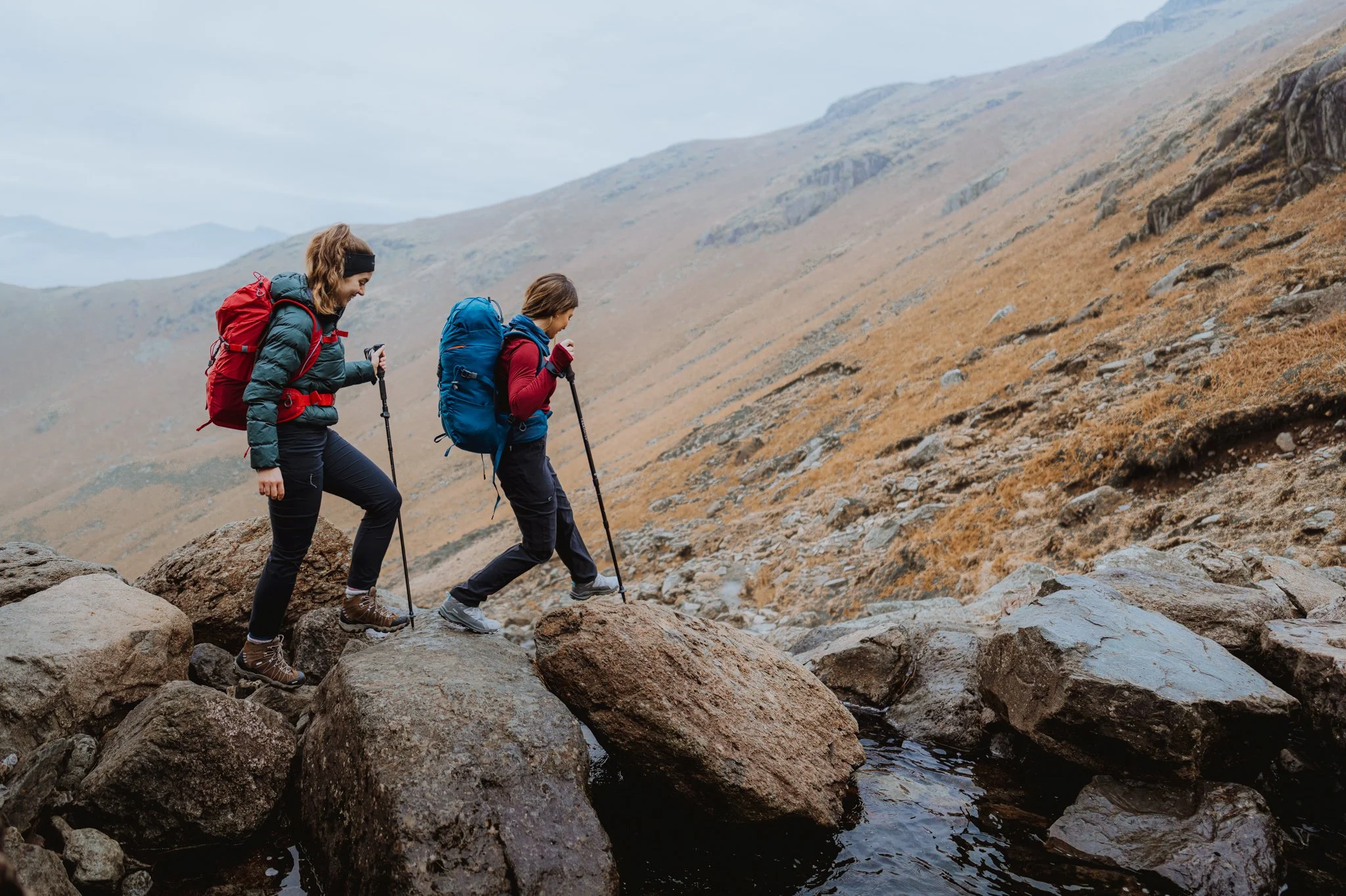 Freelance outdoor Lifestyle Photographer Two women hiking across rocks near a mountain stream, carrying backpacks and using trekking poles.