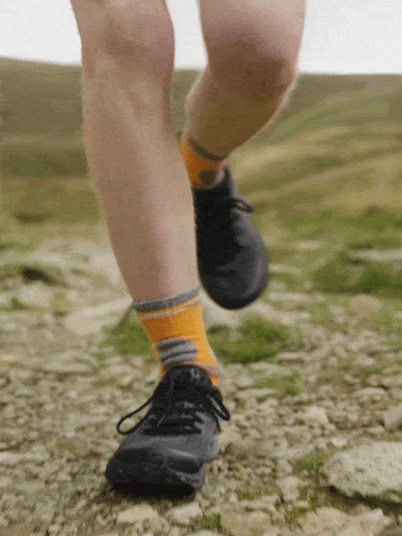 Close-up of a person running outdoors, focusing on their legs and feet, wearing black running shoes and orange socks with gray stripes, on a rocky trail.
