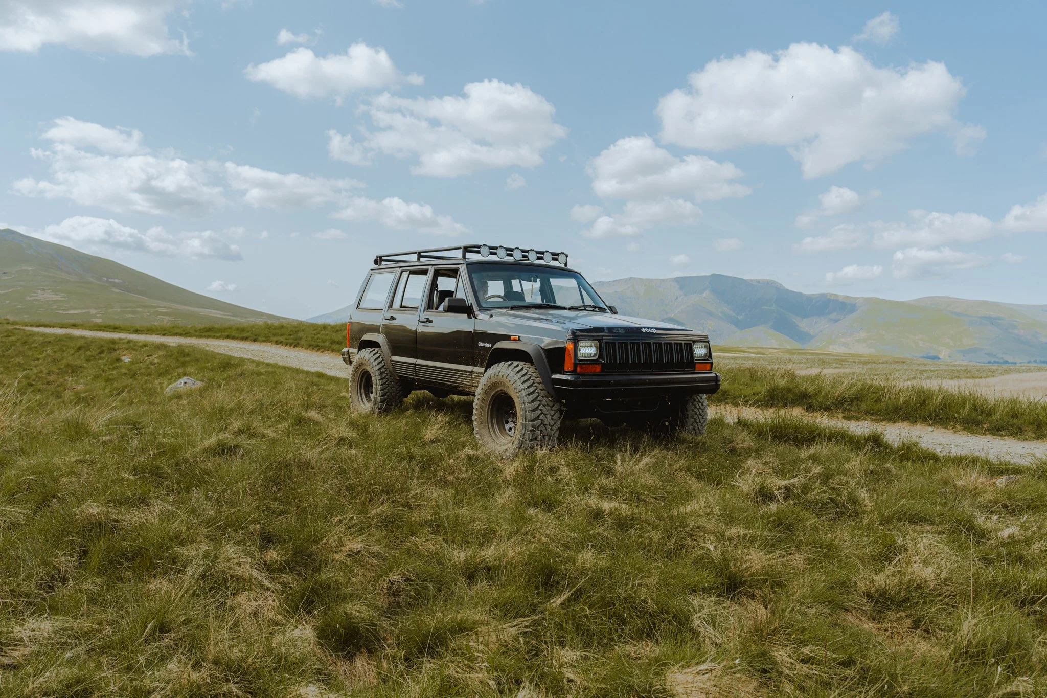 A black Jeep Cherokee with a roof rack parked on a grassy field with mountains in the background and a cloudy sky.