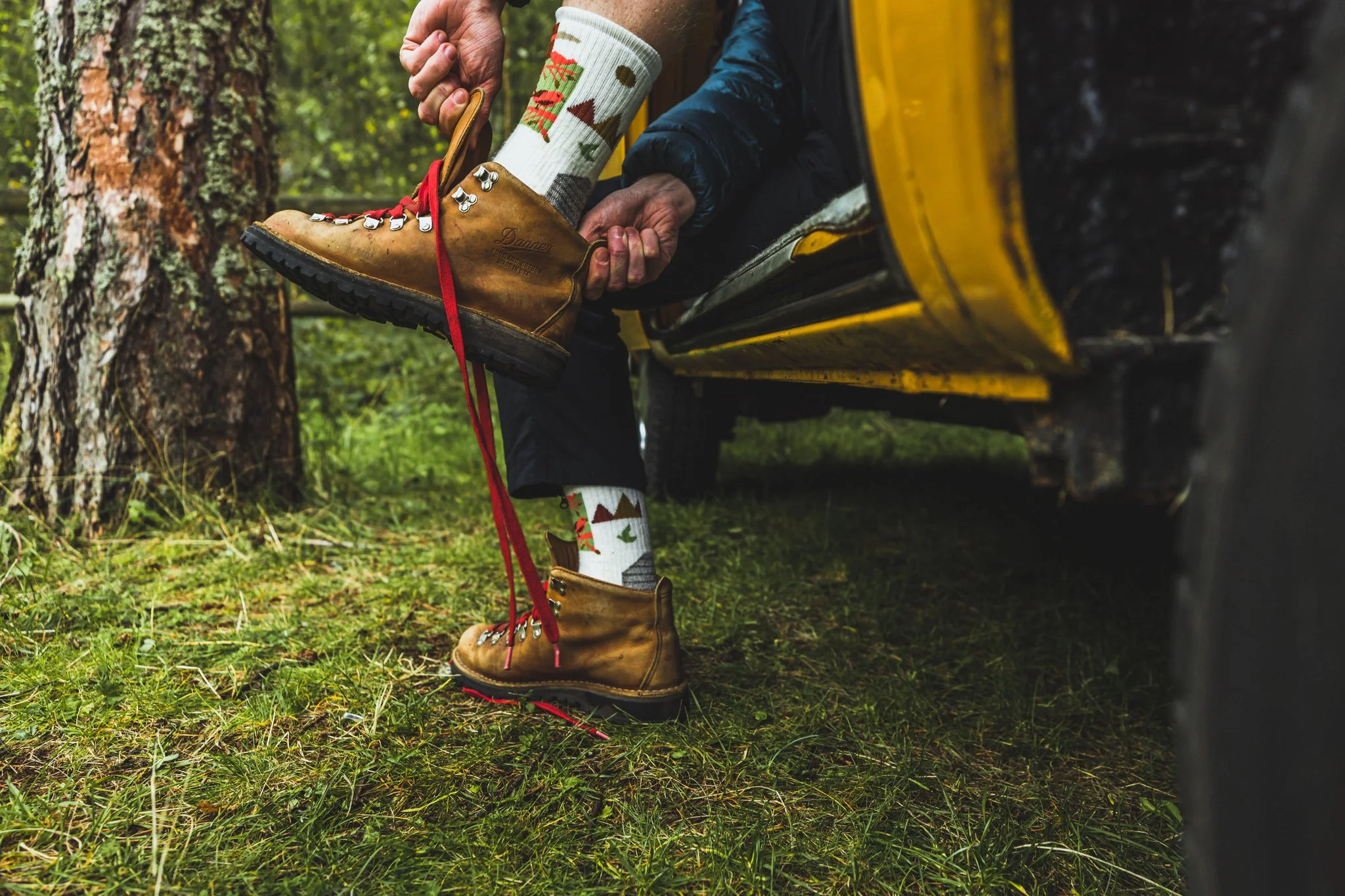 Person adjusting hiking boots while sitting on a yellow vehicle in a wooded area.