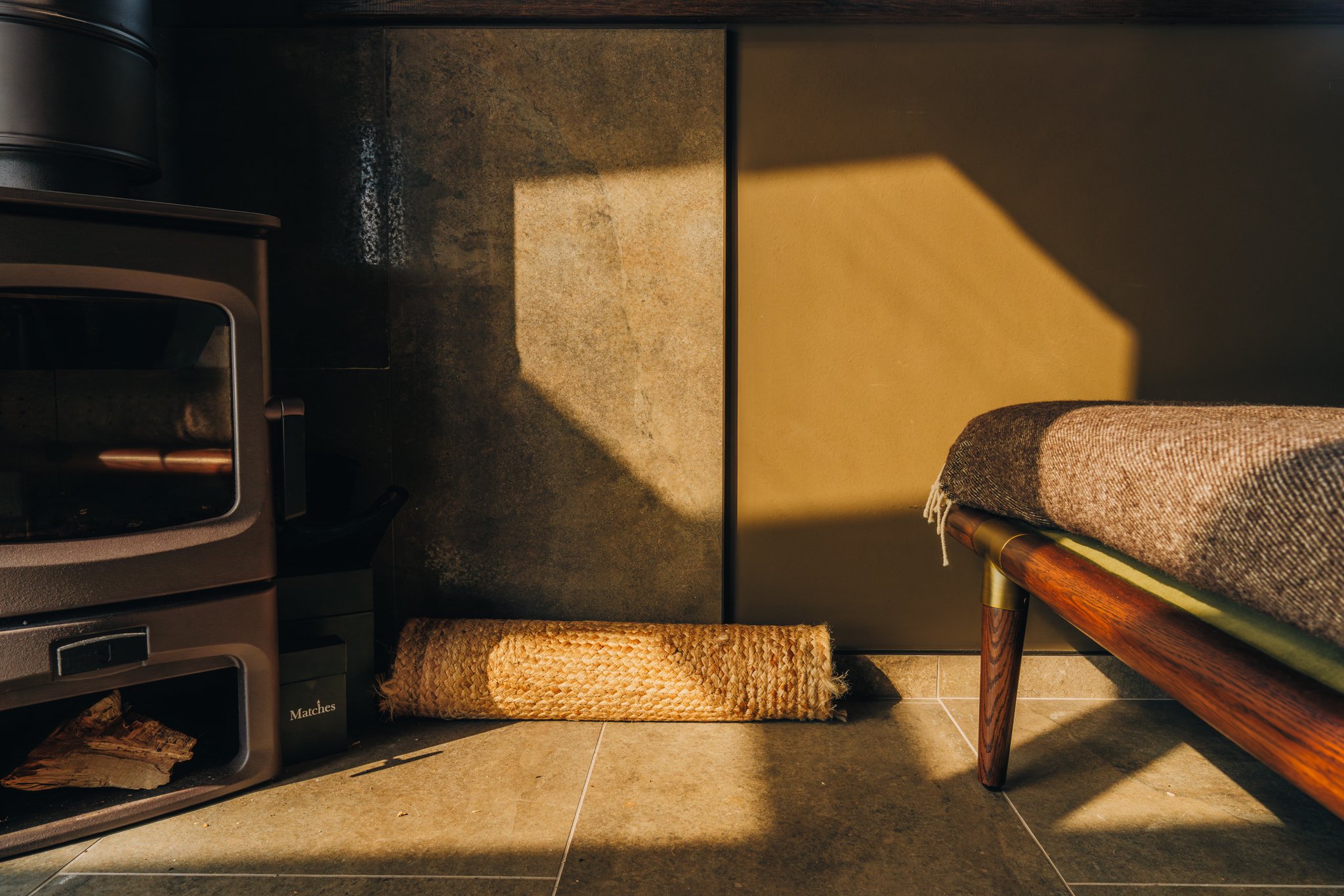 Freelance outdoor Lifestyle Photographer A cozy corner with a wood stove, a rolled rope mat, and a bench with a blanket, illuminated by warm sunlight creating a shadow on the wall.