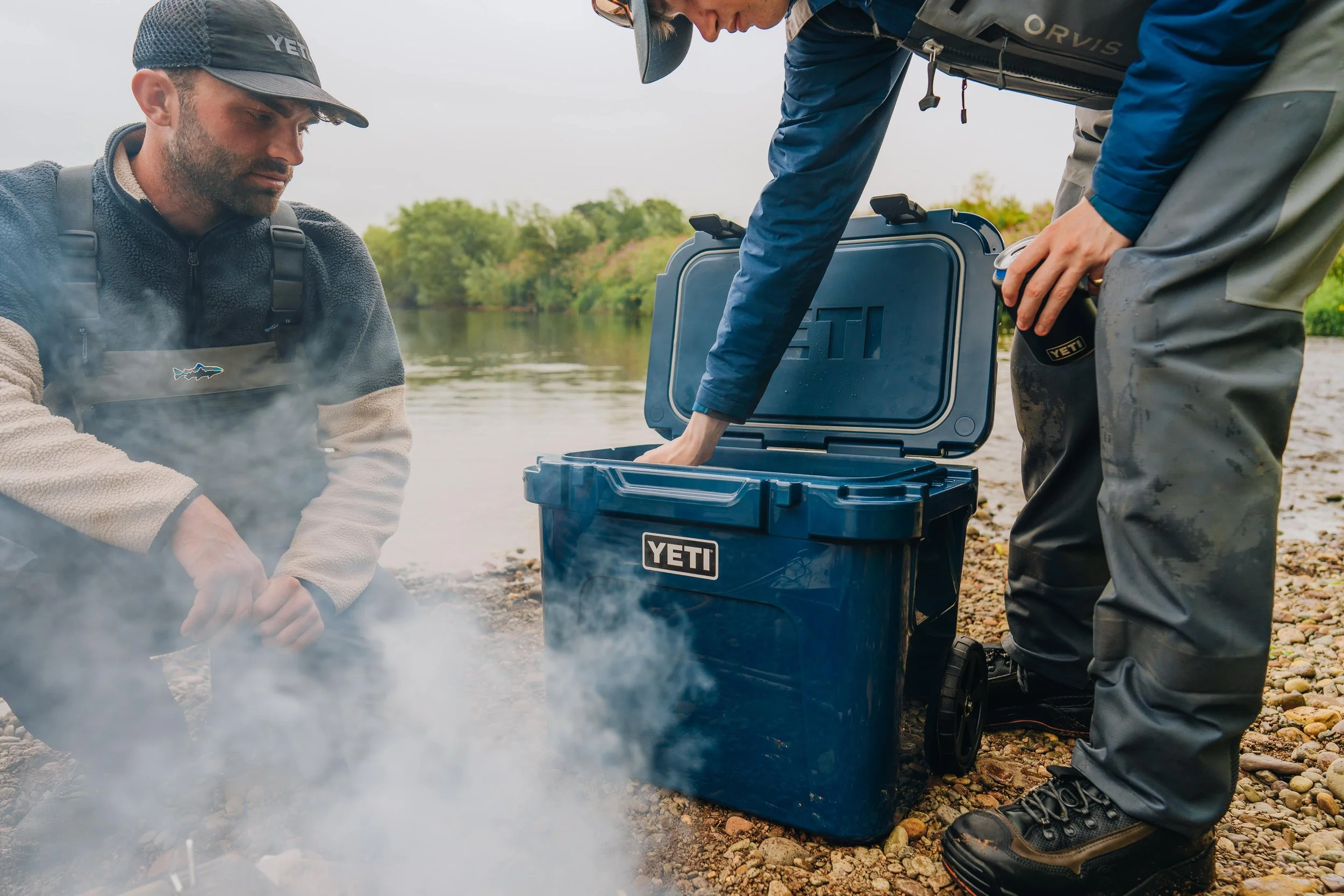 Two men are near a body of water with one of them sitting by a small fire and the other reaching into a blue YETI cooler placed on the rocky shoreline. One man is on his knees, wearing a YETI cap, beige sweater, and gray vest, while the other is bend