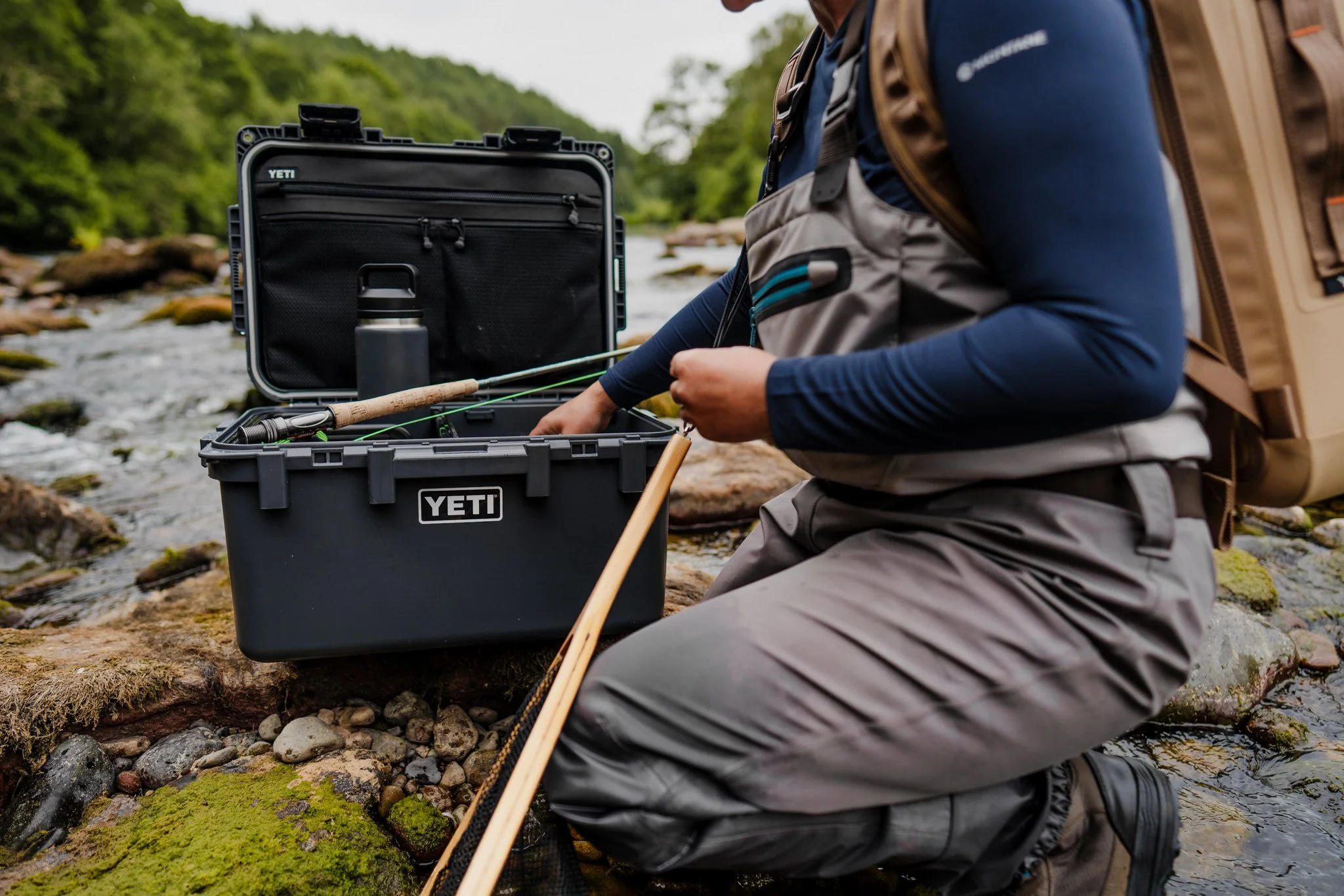 Person sitting on rocks by a river, organizing fishing gear from a YETI cooler, with fishing rod, water bottle, and outdoor clothing. Freelance Lifestyle Photographer