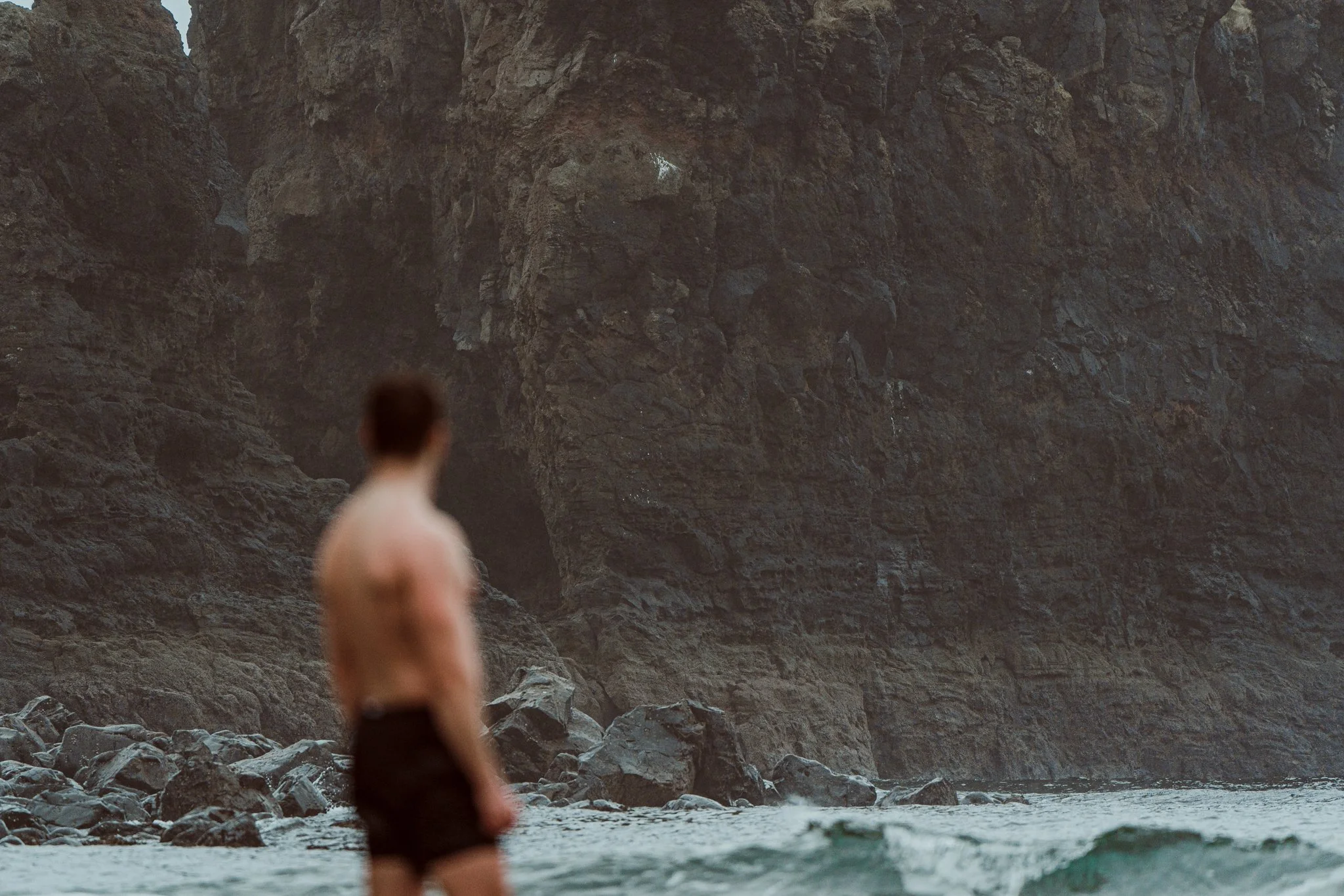 A person standing in shallow water near a rocky shoreline with large cliffs in the background.