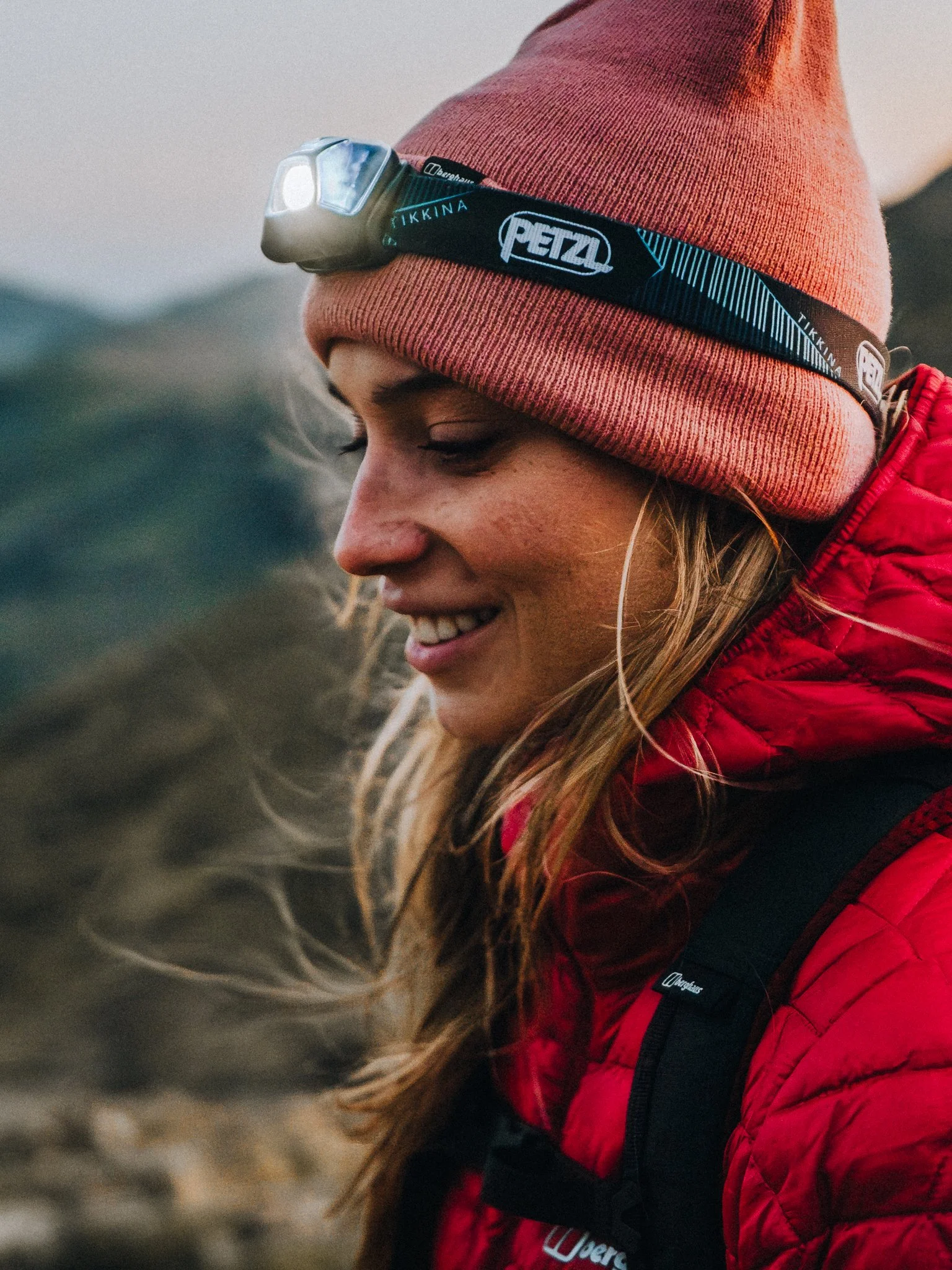 A person in a red jacket and a red knit hat with a headlamp on it, with long hair, smiling and looking down.