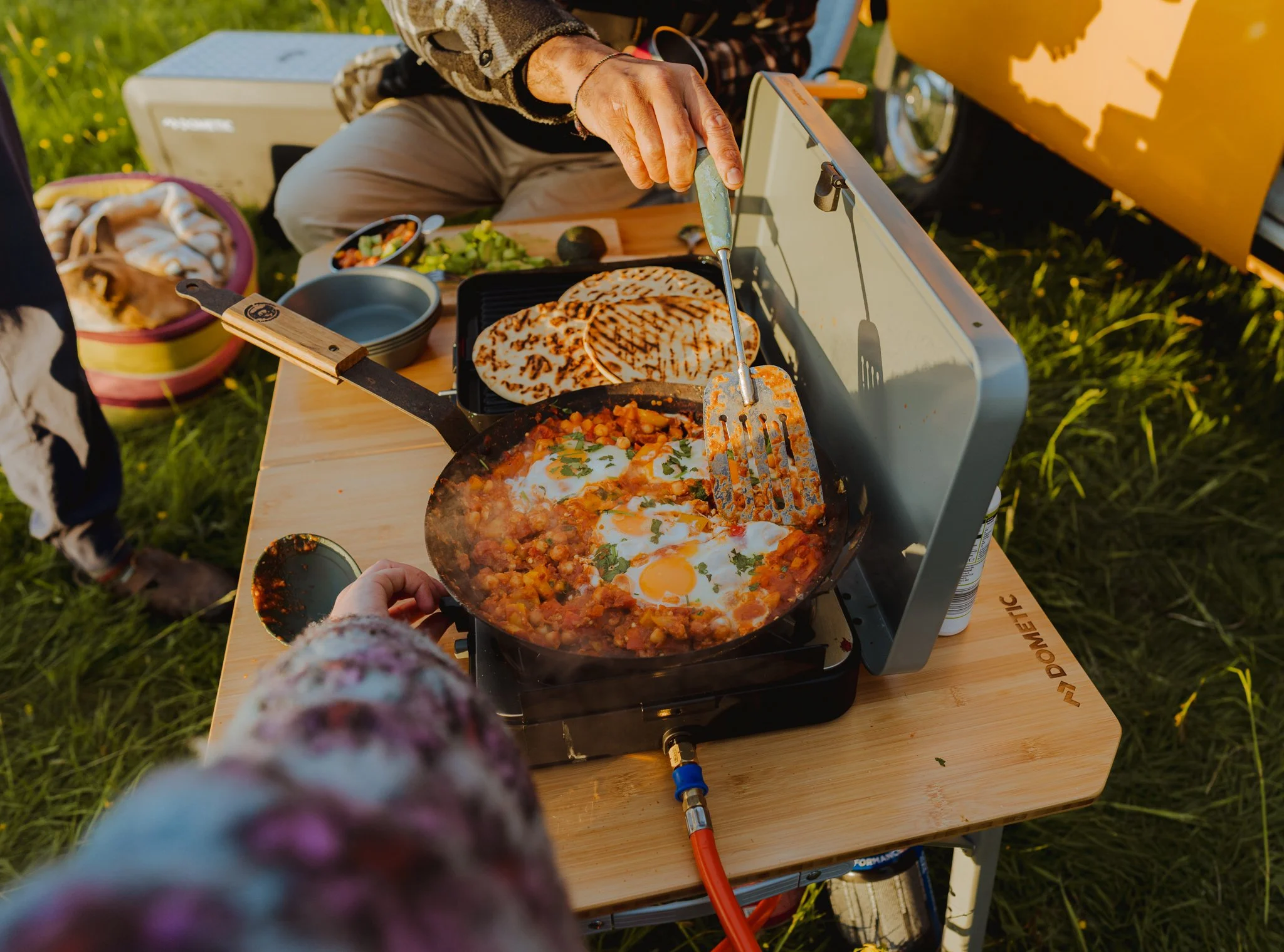 Person adding eggs to a skillet with cooked ground meat and tomato sauce on an outdoor camping stove, with grilled bread and a bowl of vegetables on a wooden table.