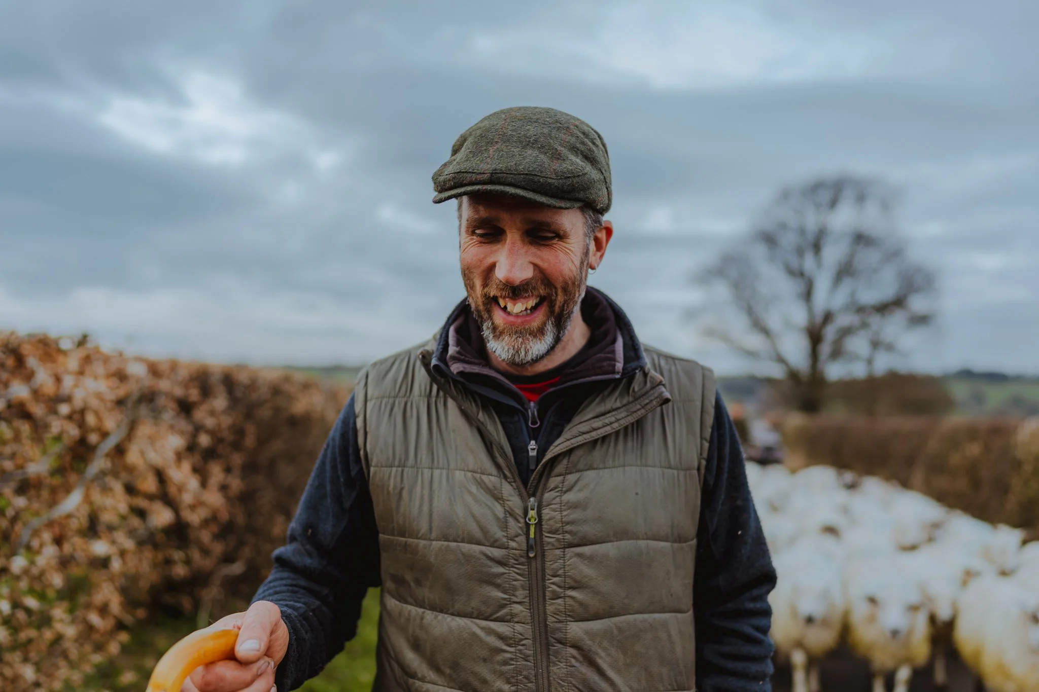 A man smiling outdoors holding a yellow tool, with sheep and trees in the background on a cloudy day.