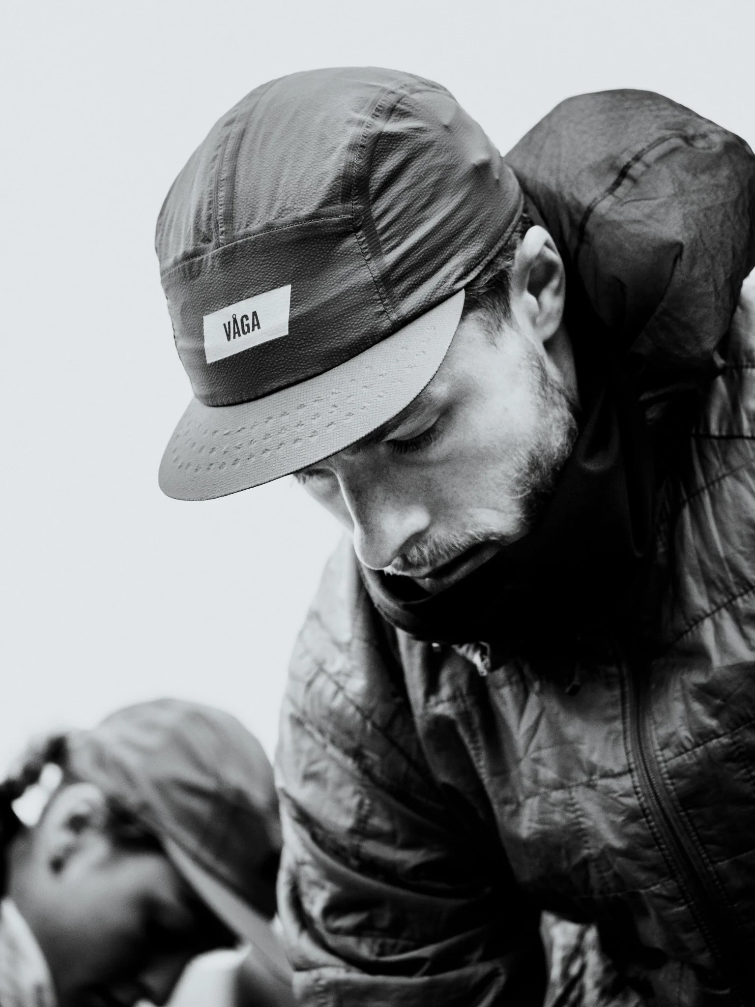 Freelance outdoor Lifestyle Photographer Black and white photo of a man wearing a cap with the word "VAGA" on it, looking downward with another person in the background.