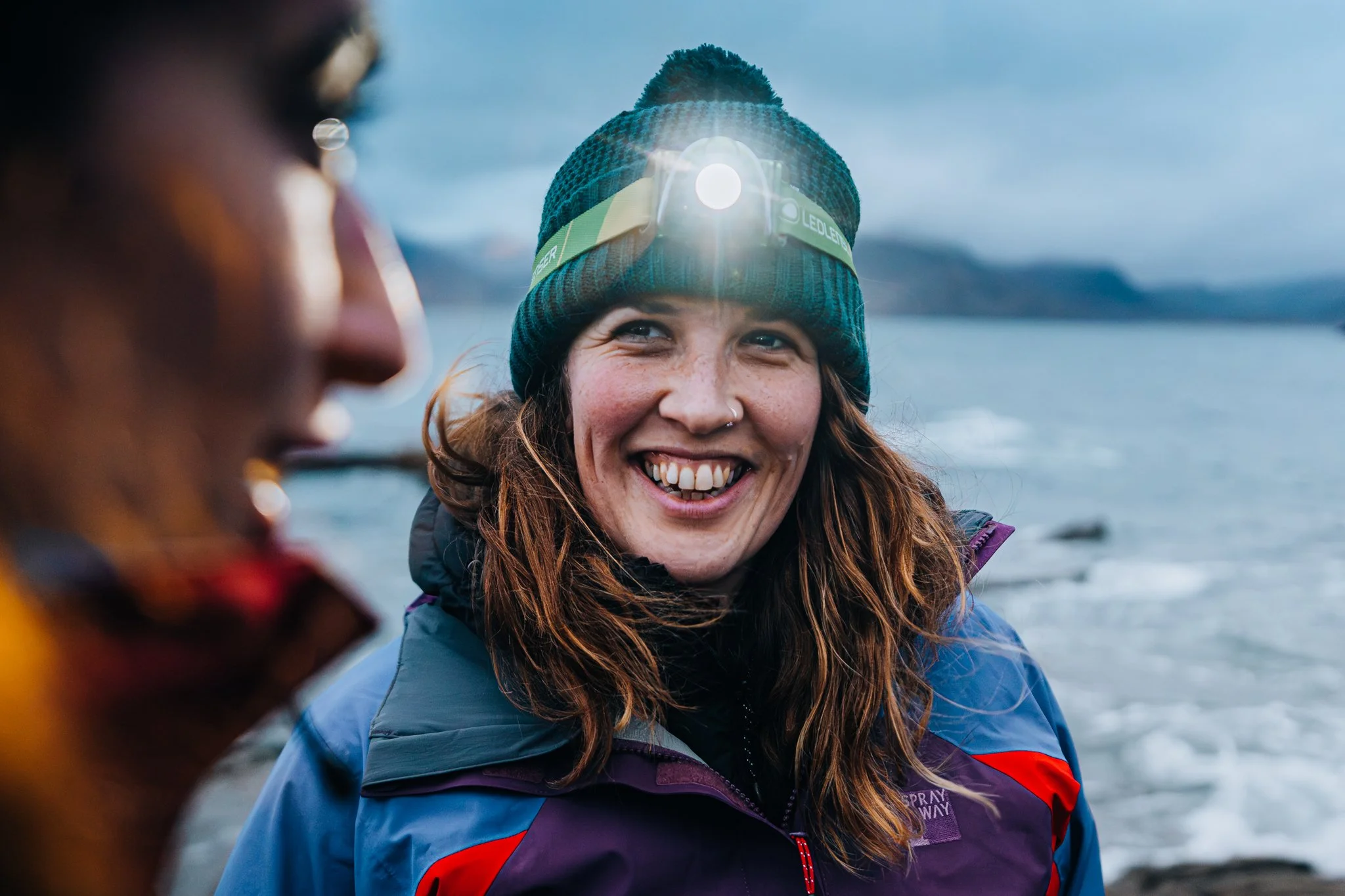 Freelance Lifestyle Photographer A smiling woman wearing a knit beanie with a headlamp, a blue and purple jacket, standing near the ocean with mountains in the background.