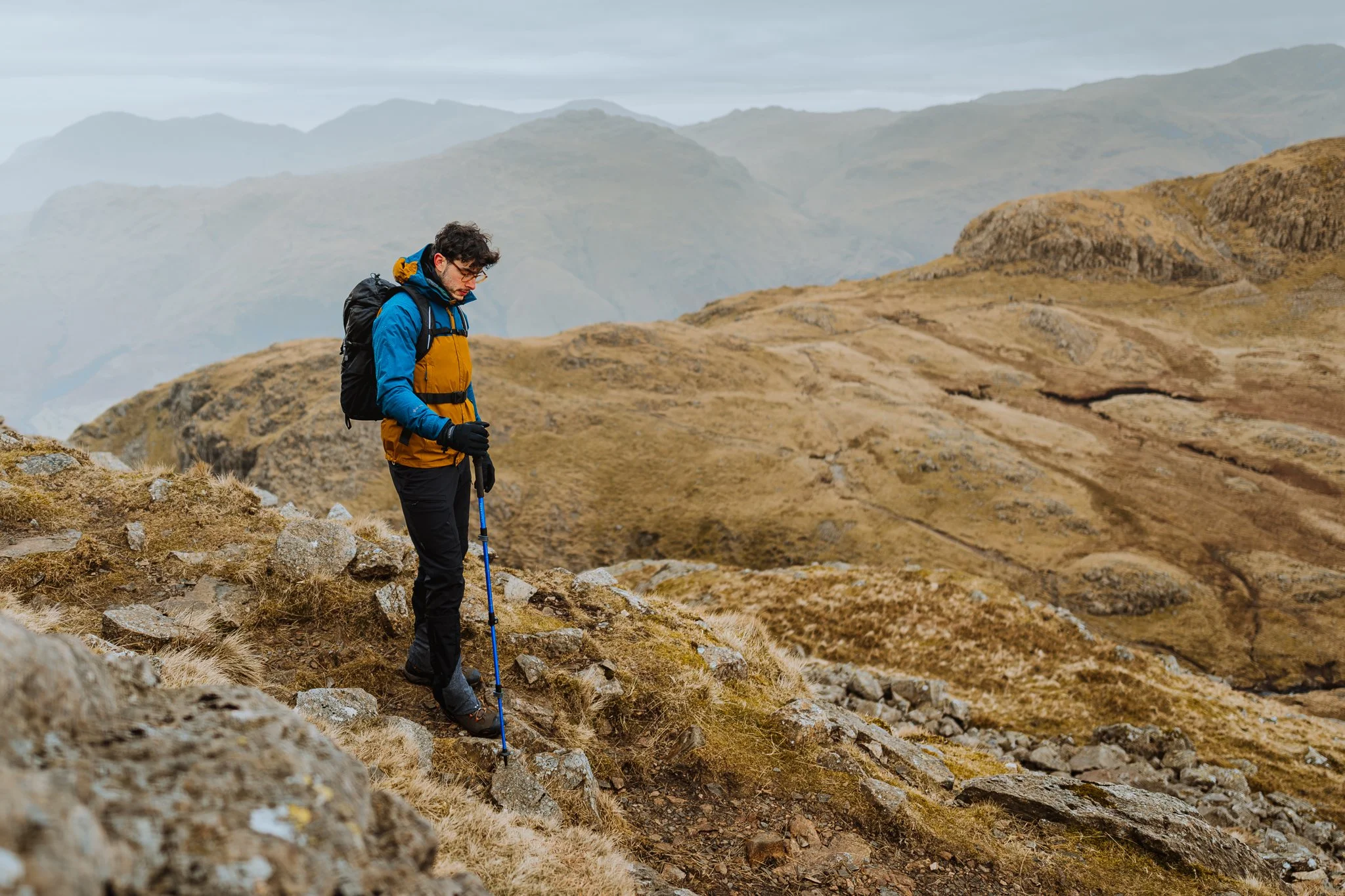 Freelance outdoor Lifestyle Photographer A man hiking down a rocky trail in a mountainous landscape during overcast weather, wearing a blue and brown jacket, black pants, gloves, and carrying a backpack and hiking poles.