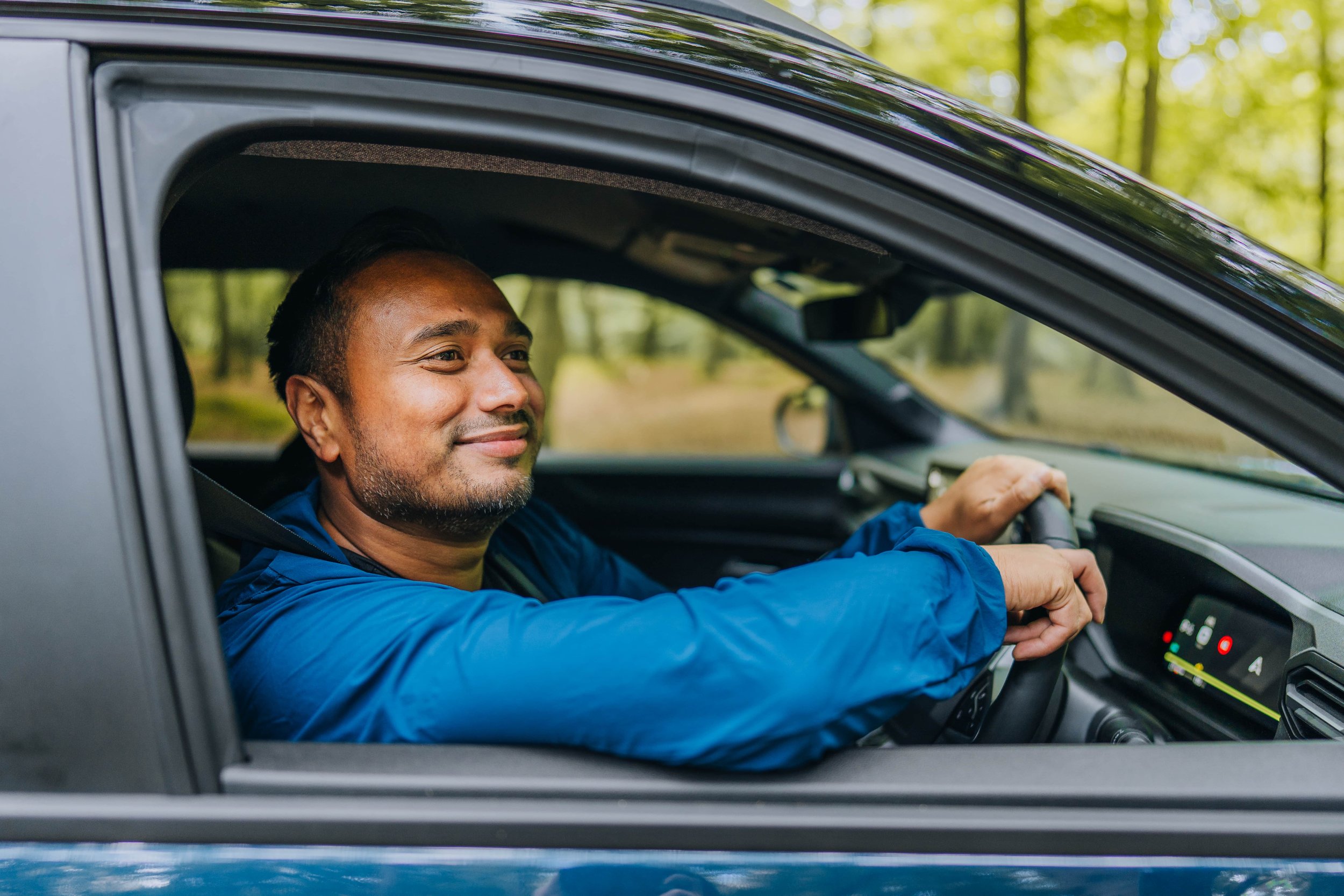 A man with a beard sitting in the driver's seat of a car, smiling, with trees and greenery visible outside the window.