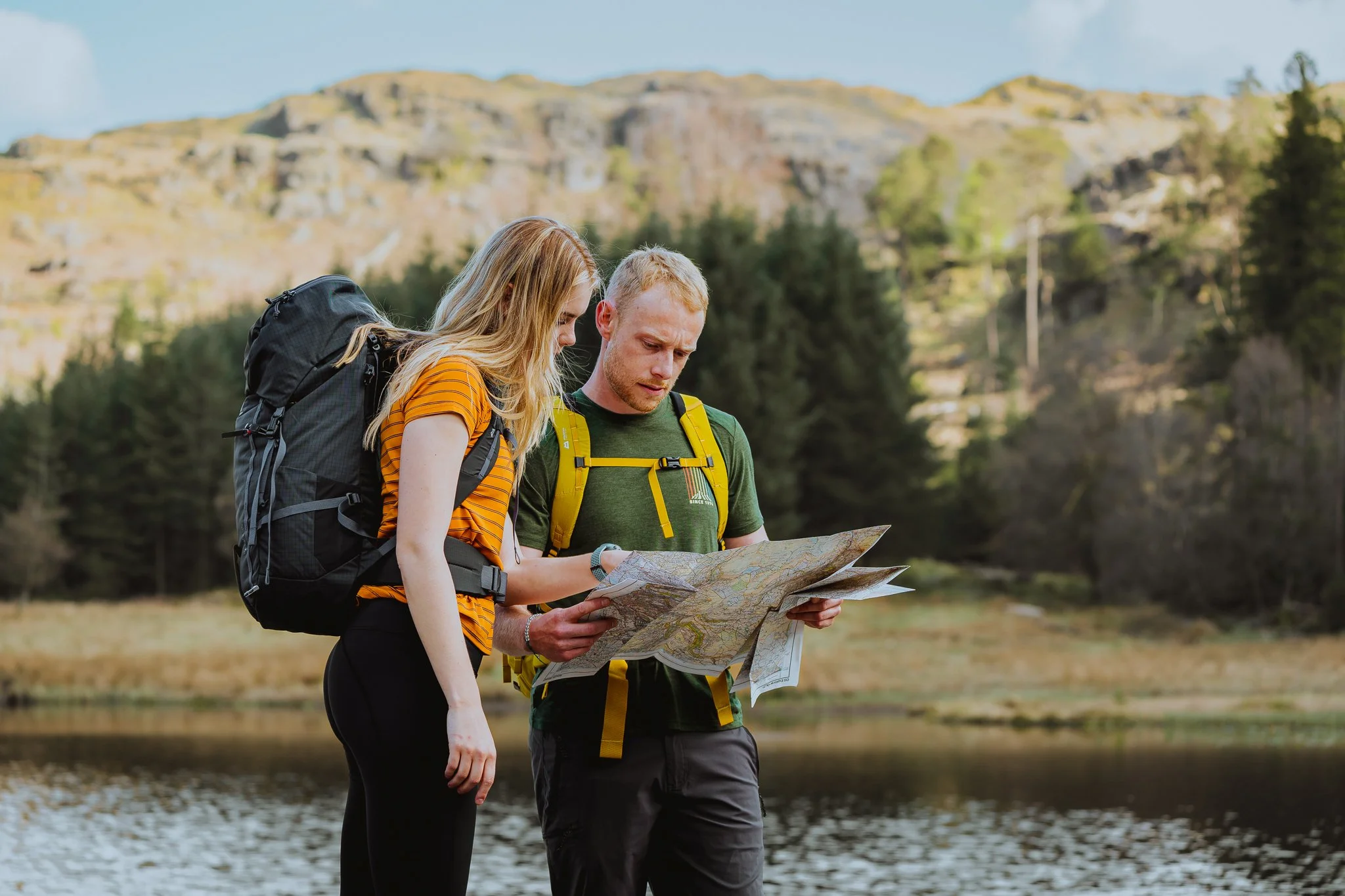 A man and a woman looking at a map while hiking near a lake with trees and hills in the background.