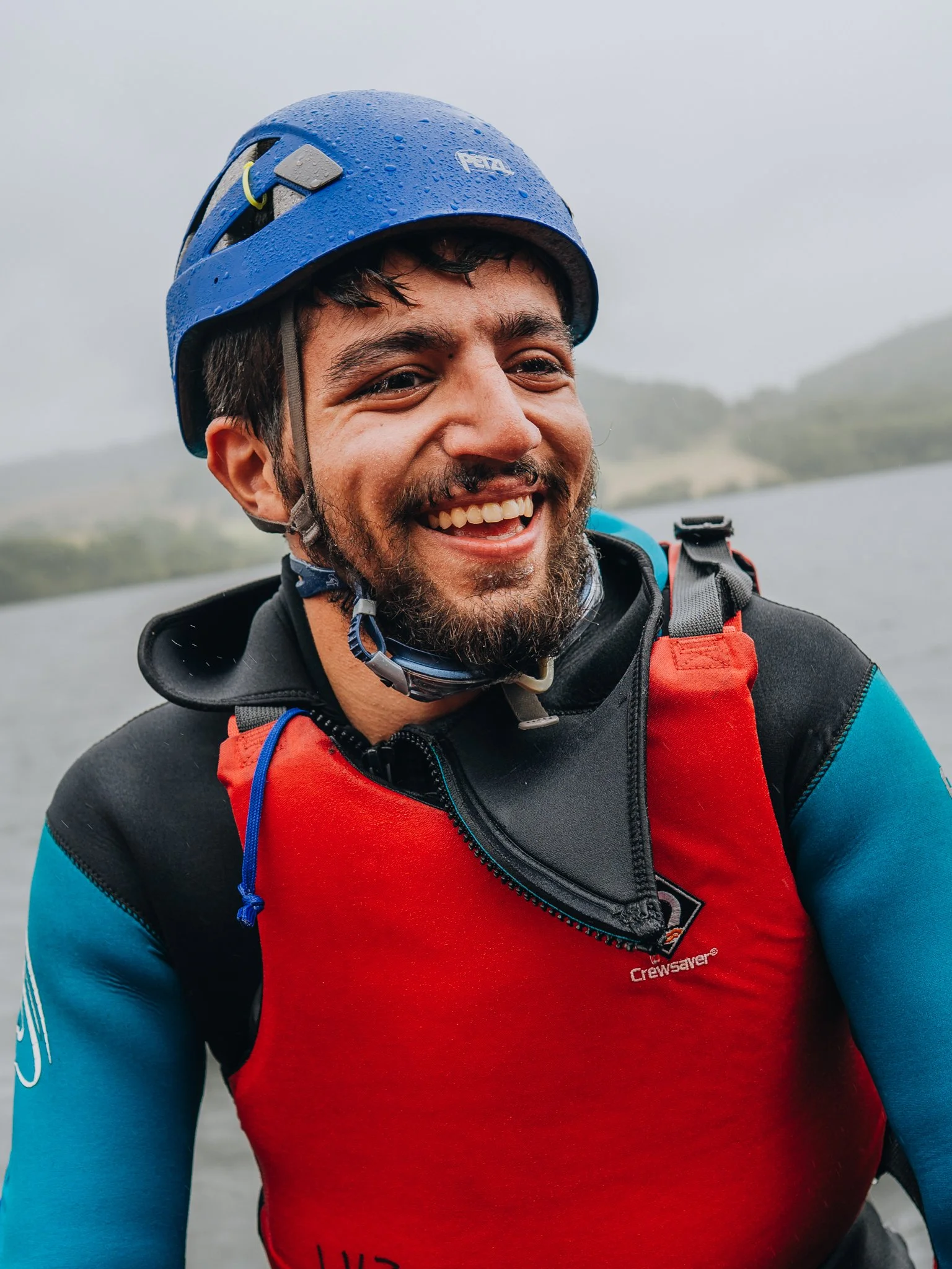 Freelance Lifestyle Photographer A smiling man wearing a blue helmet and a red life jacket outdoors near water, with cloudy sky and distant mountains in the background.