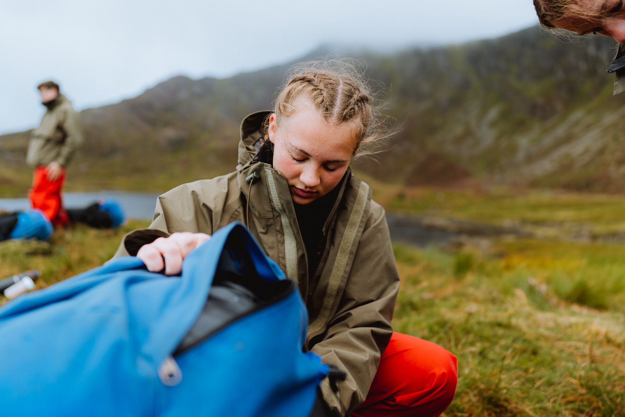 Freelance outdoor Lifestyle Photographer A young girl packing a blue backpack outdoors in a scenic natural landscape with hills and a body of water, possibly on a camping trip.