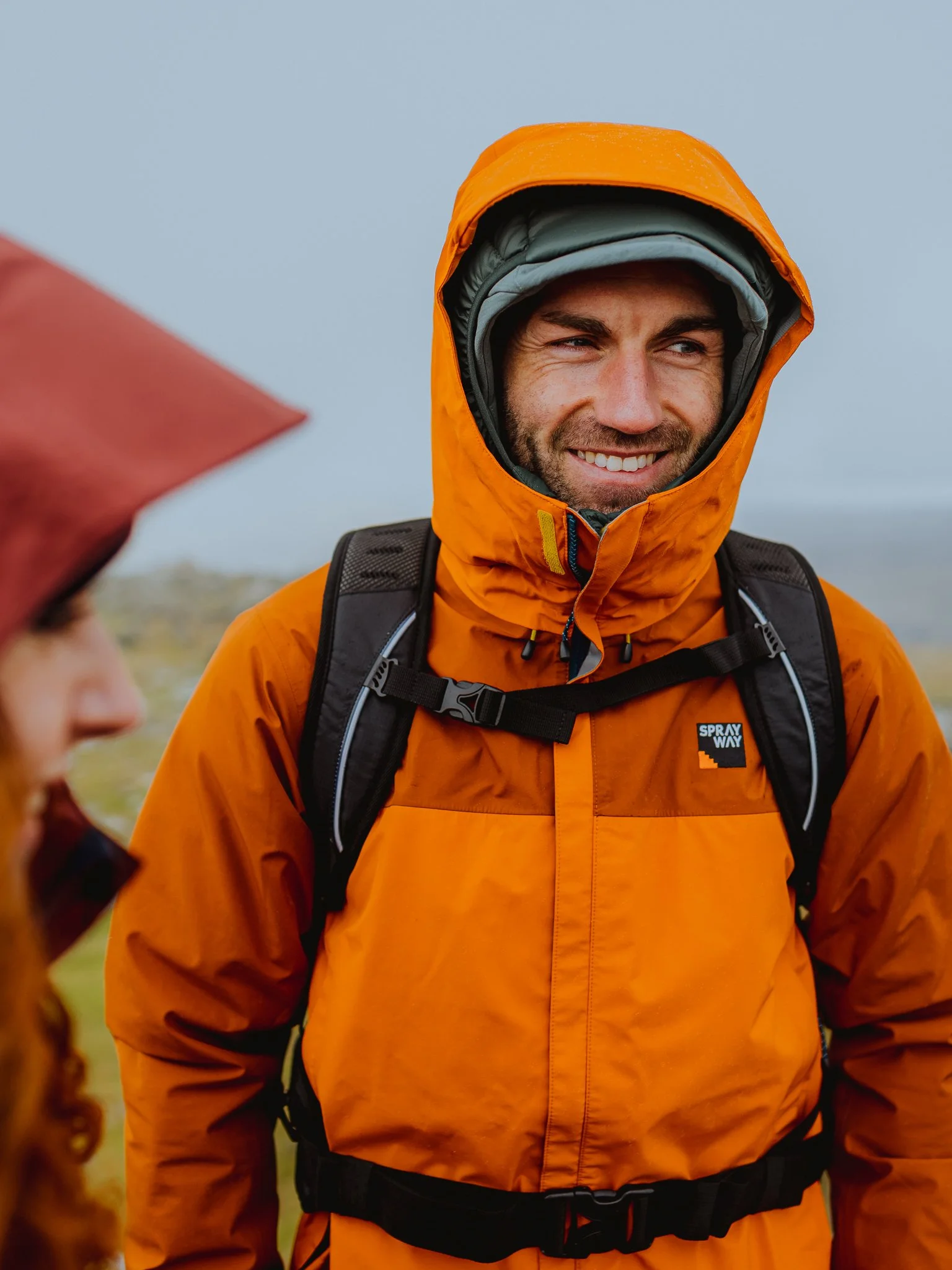 Freelance Lifestyle Photographer A man smiling outdoors wearing an orange rain jacket with a hood and a backpack, with a woman partially visible in the foreground, also dressed for outdoor activity.
