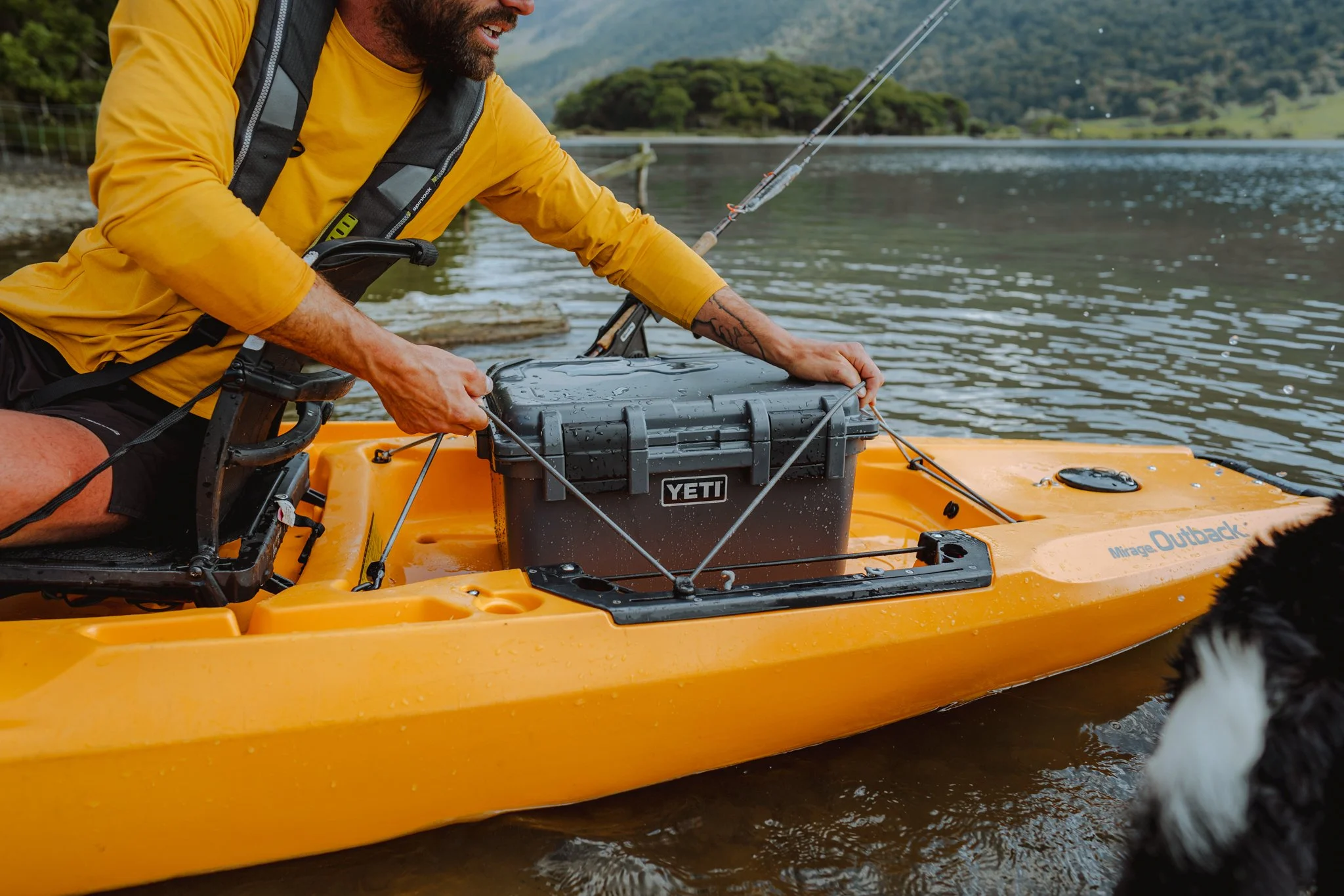 A man in a yellow shirt on a yellow kayak named 'Mirage Outback' is placing a black YETI cooler in the kayak. There are mountains and a lake in the background, and a black and white border collie dog is partially visible in the water near the kayak.