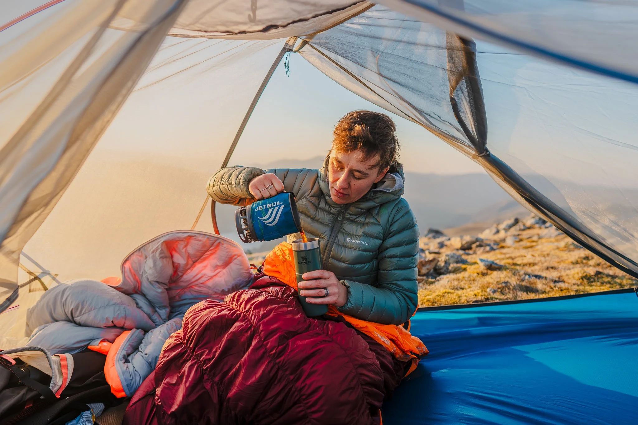 A person inside a tent pouring hot beverage into a cup, surrounded by sleeping bags, with rocky terrain outside.