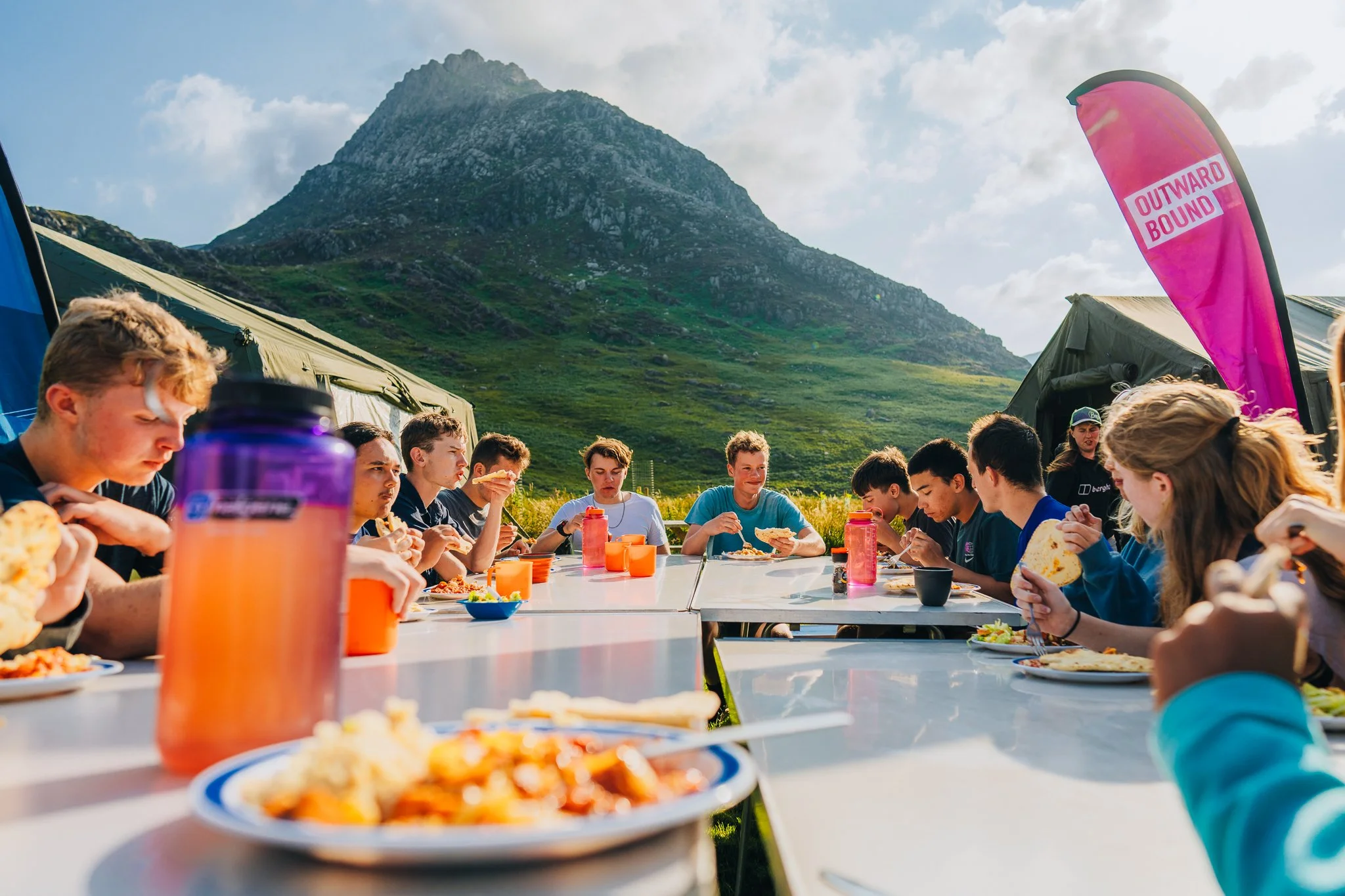Group of teenagers sitting at a long outdoor table enjoying a meal during a camping trip in a scenic mountain area, with tents and a pink flag in the background.