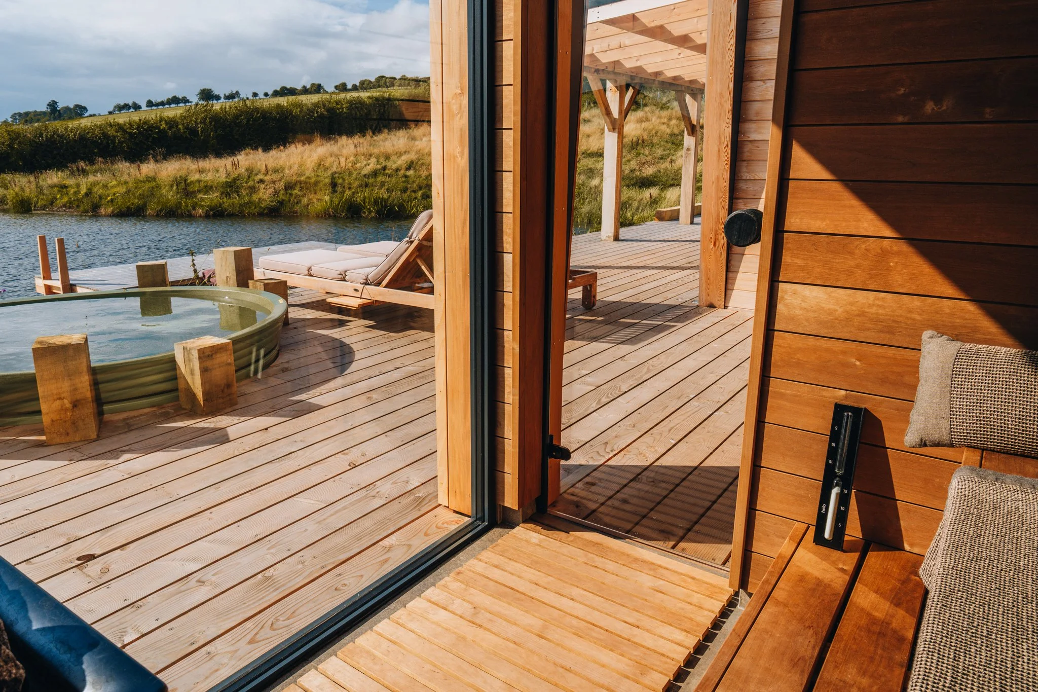 Freelance outdoor Lifestyle Photographer View from inside a wooden sauna looking out onto a deck with a hot tub, lounge chairs, and a scenic river and grassy hills in the background.
