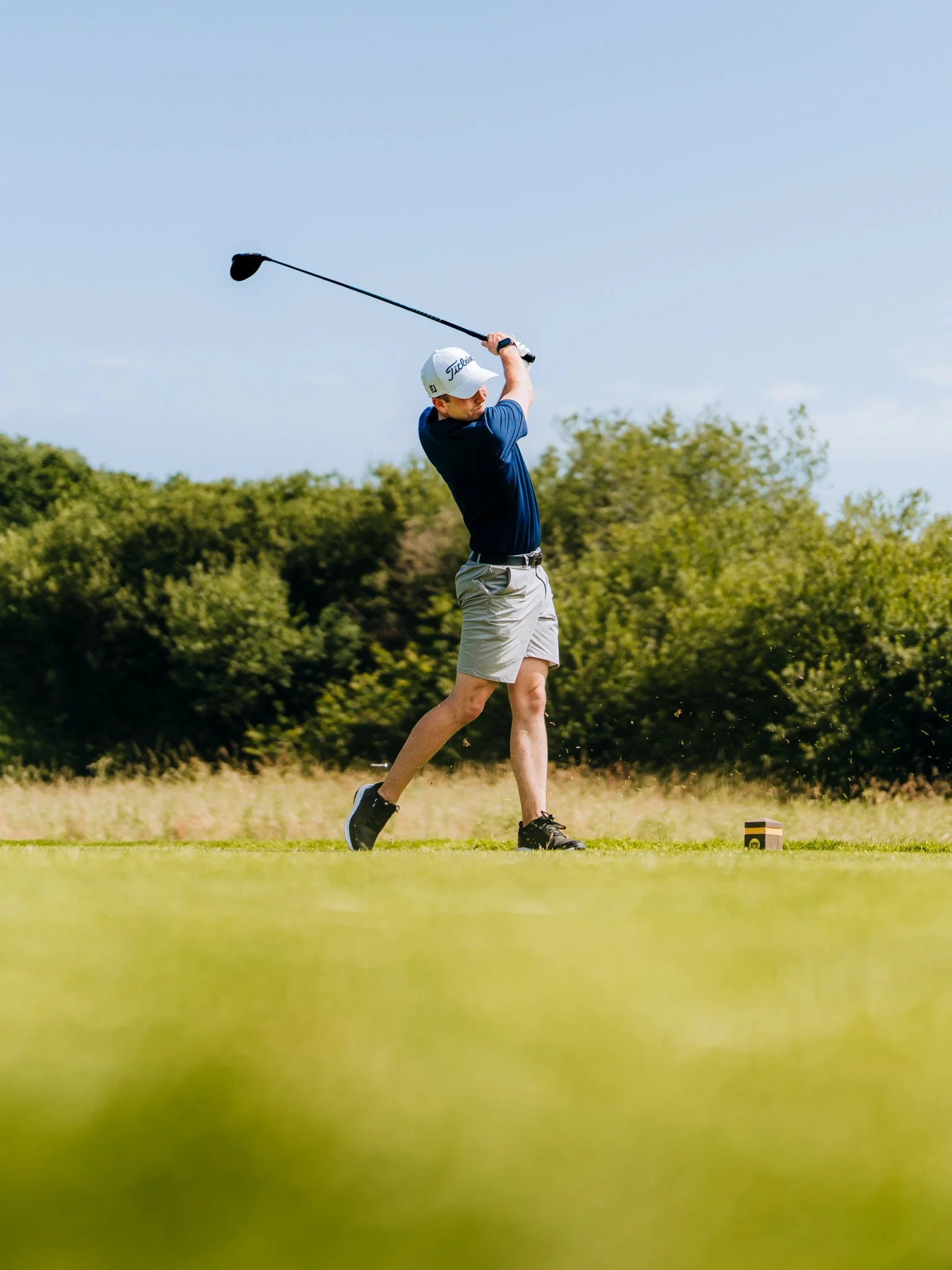 A man playing golf on a course, swinging a club with a blue sky and green trees in the background.
