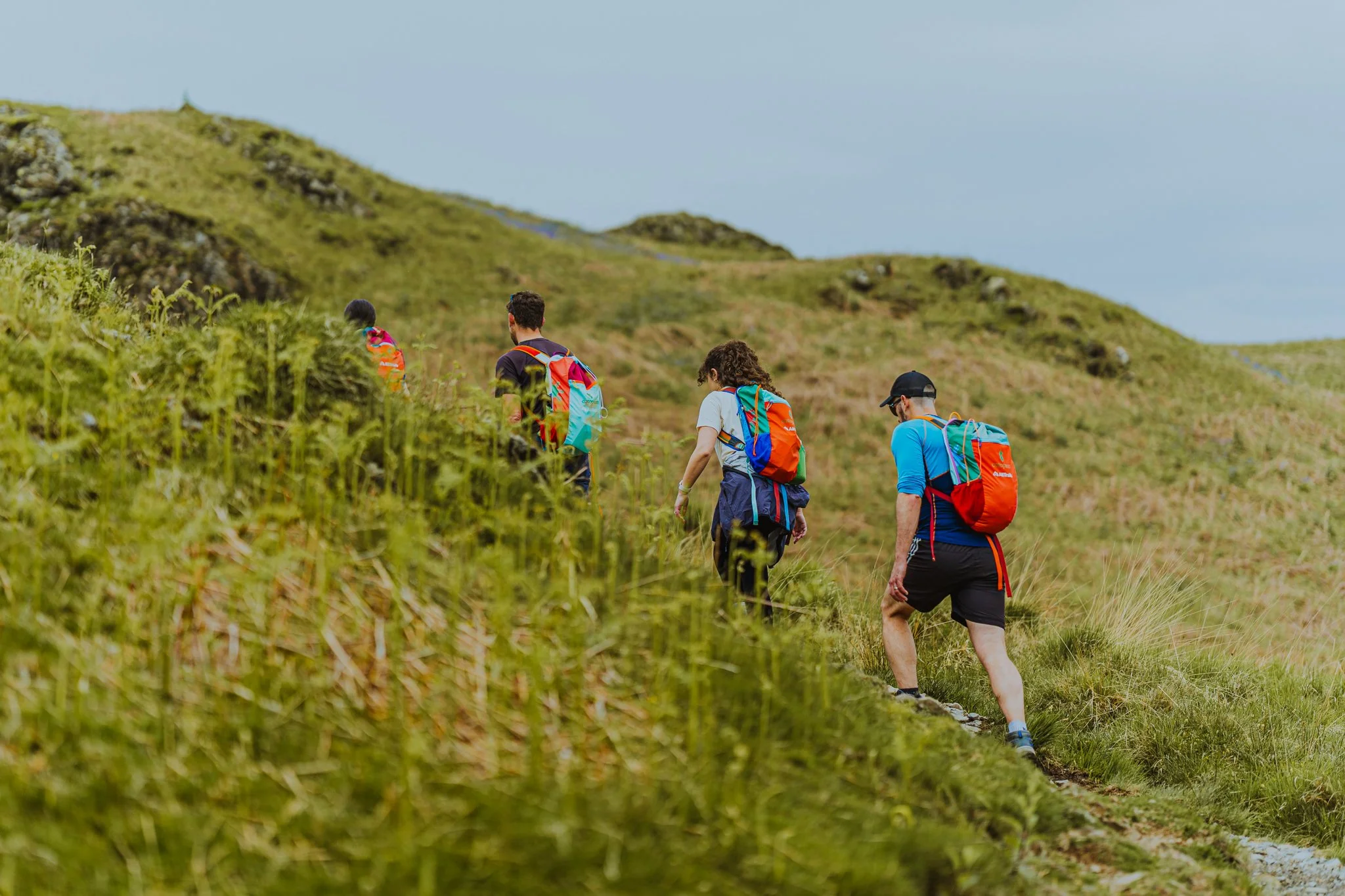 Freelance outdoor Lifestyle Photographer Four hikers with backpacks walking along a trail through green grassy hills and mountains under a cloudy sky.