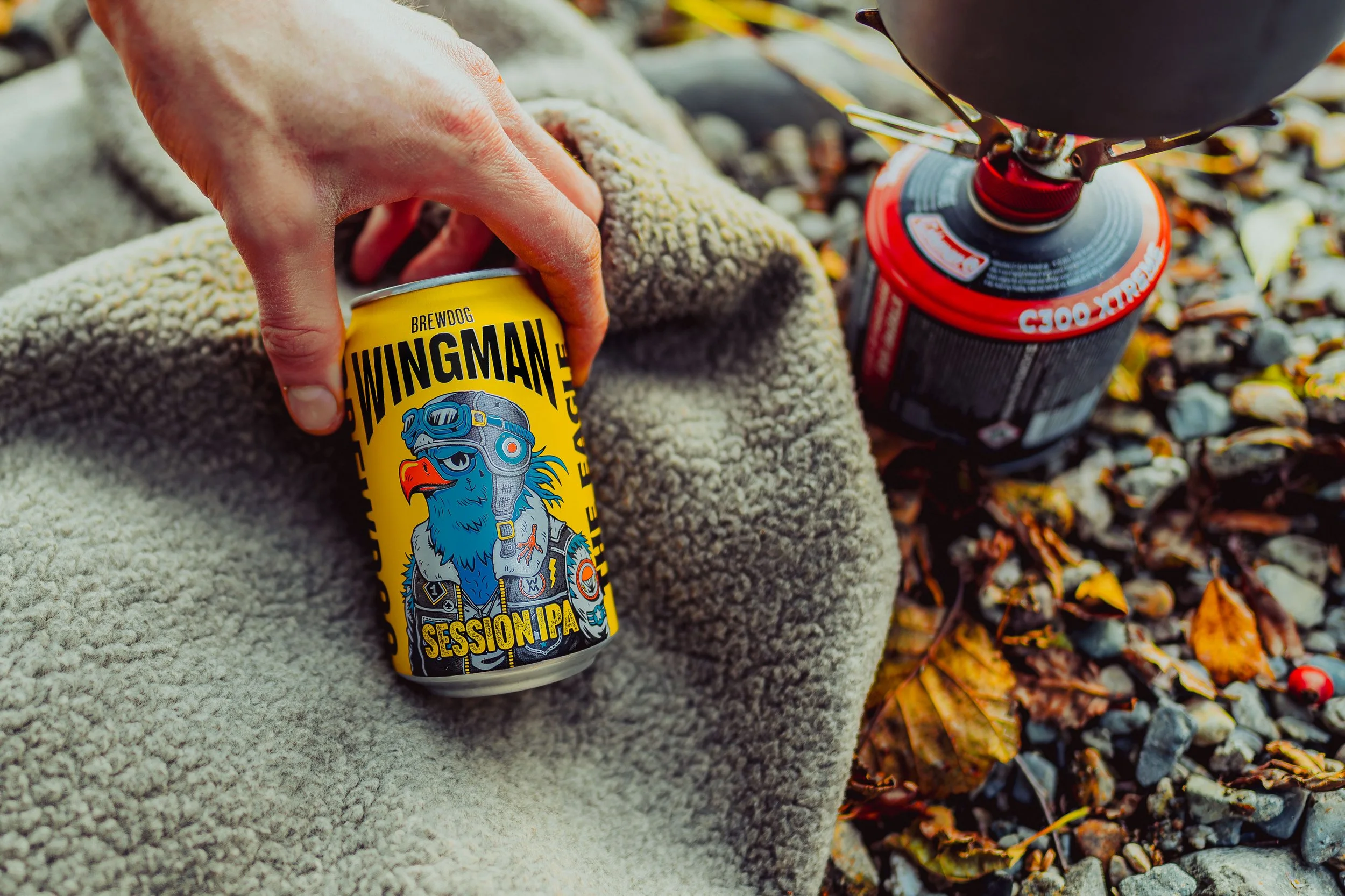 Freelance Lifestyle Photographer A person is sitting on a blanket or towel on the ground covered with fallen autumn leaves and small rocks, holding a yellow can of Brewdog Wingman Session IPA beer. A red camping stove is nearby.